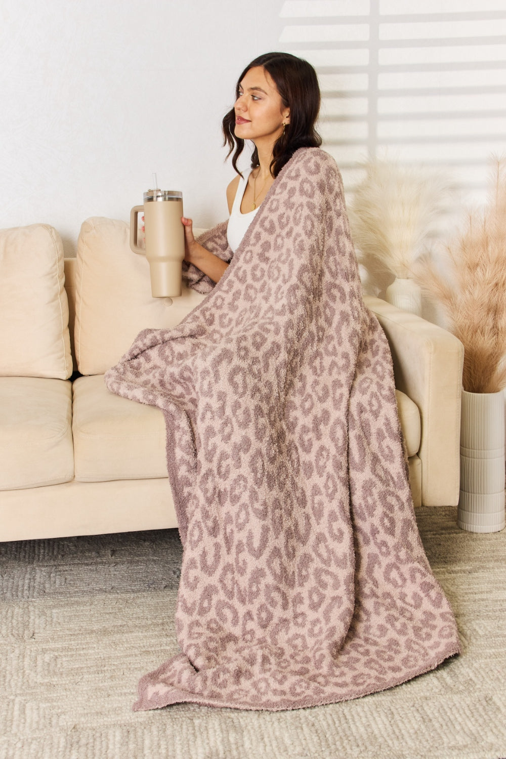 A woman stands in a room holding up the Cuddley Leopard Decorative Throw Blanket, which features a large, leopard-print design in a neutral color scheme. She is smiling and facing the camera, showcasing the 100% polyester fabric.