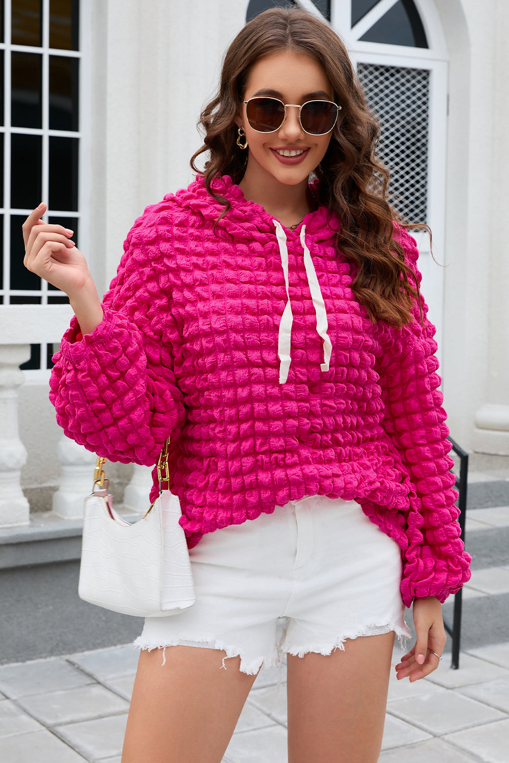 A person stands with their back to the camera, donning the Rose Bubble Textured Waffle Hoodie in a vibrant pink hue. They are in a room with yellow floral wallpaper, and a corner of a wooden chair is visible.