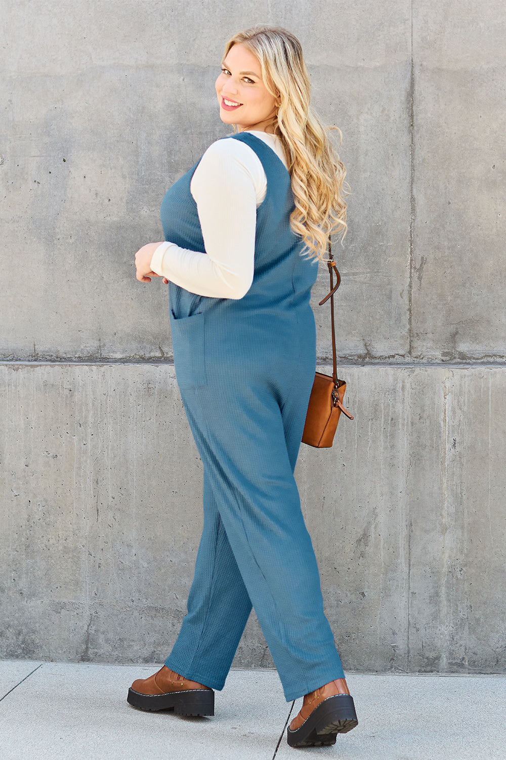 A person stands outdoors wearing the Double Take Full Size Sleeveless Straight Jumpsuit in blue and brown sandals, holding a cream-colored bag. In the background, there's a wooden bench and palm trees.