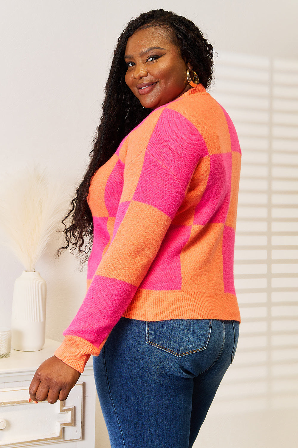 A woman with long hair stands smiling, wearing the Angel Wings Woven Right Checkered V-Neck Dropped Shoulder Cardigan in pink and orange over blue jeans.