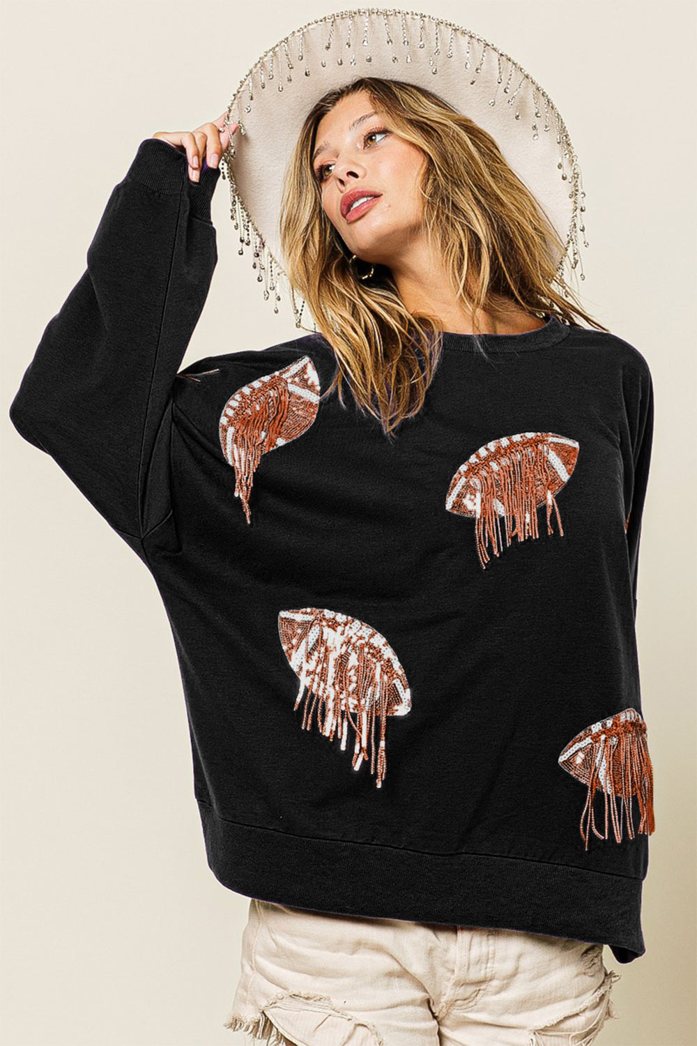 A woman dons an oversized BiBi Sequin Fringe Football Patch Round Neck Sweatshirt, adorned with jellyfish designs, while pairing it with a beige fringed hat. She strikes a pose with one hand on the brim of her hat, her hair windswept as she gazes upward, fully embracing a sporty style.