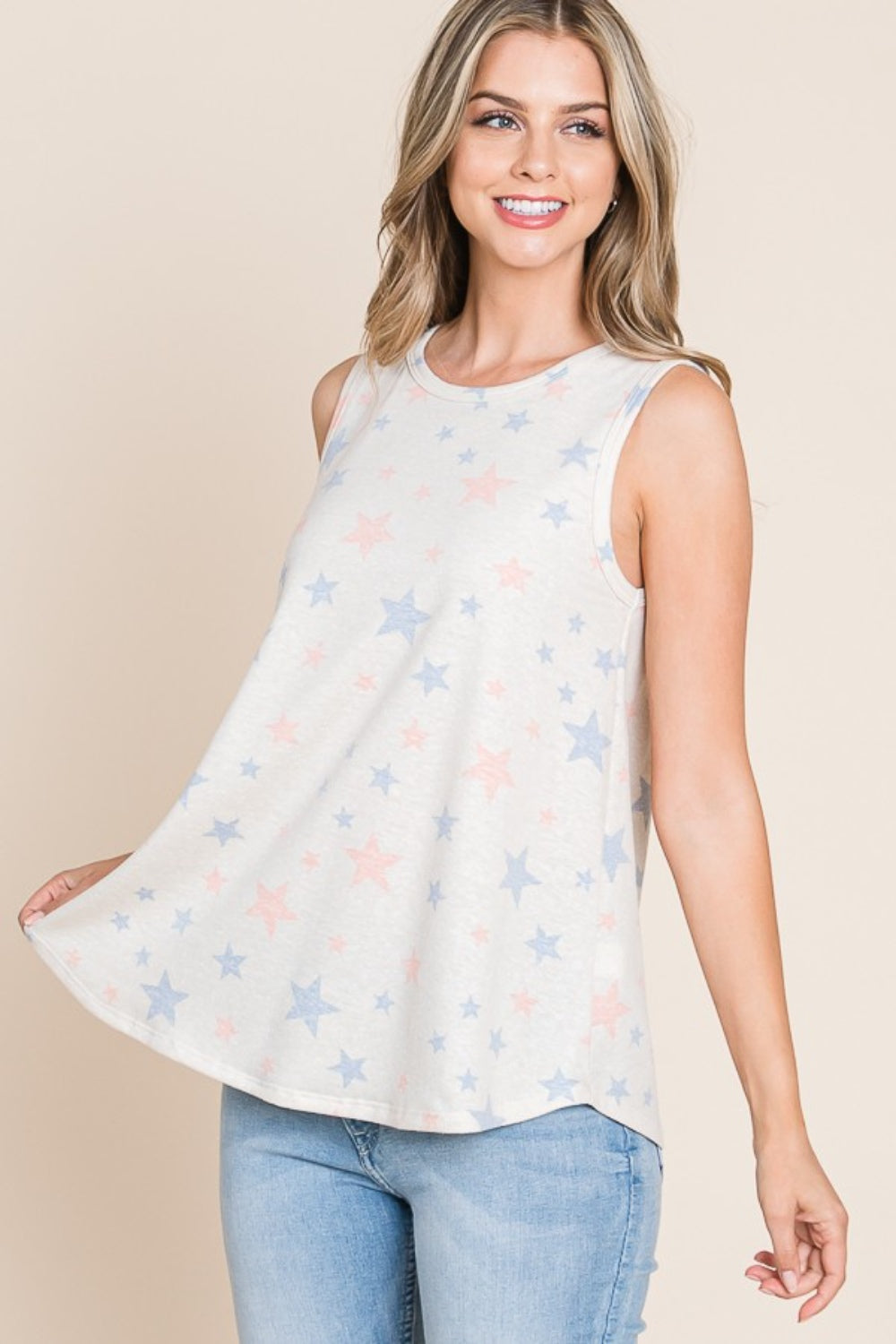 A woman with long, wavy hair is standing and smiling against a beige background, dressed in a casual wear ensemble featuring the BOMBOM Star Print Round Neck Tank with blue and pink patterns, paired with light blue jeans.