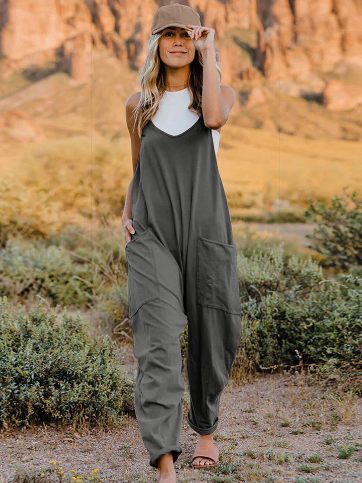 A woman wearing the Double Take Full Size Sleeveless V-Neck Pocketed Jumpsuit in green and a beige cap stands outdoors in a desert landscape with plants and rocky cliffs in the background.