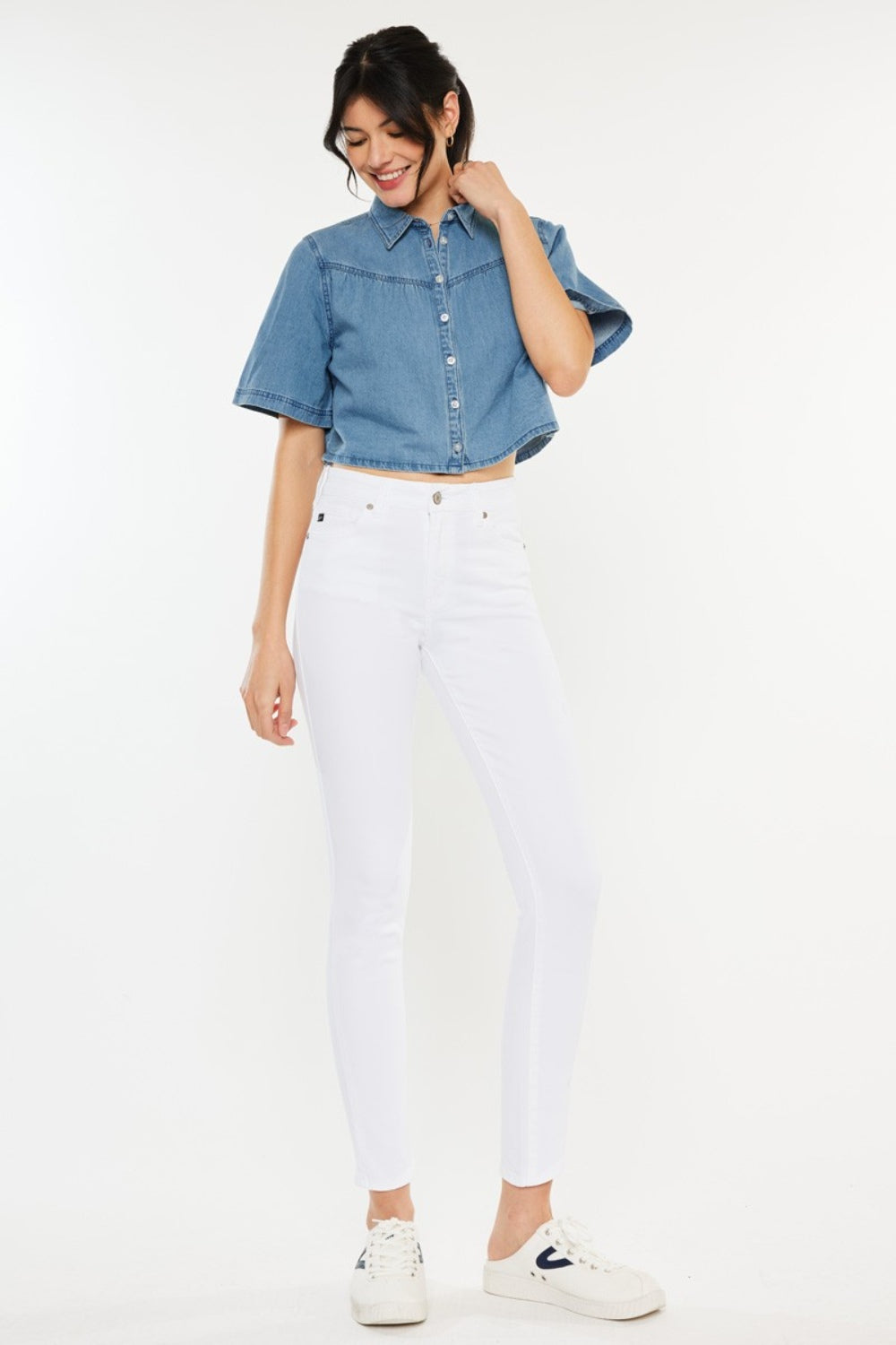 A person wearing a short-sleeve denim button-up shirt, Kancan High Rise Ankle Skinny Jeans in classic white, and white sneakers stands effortlessly against a plain background.