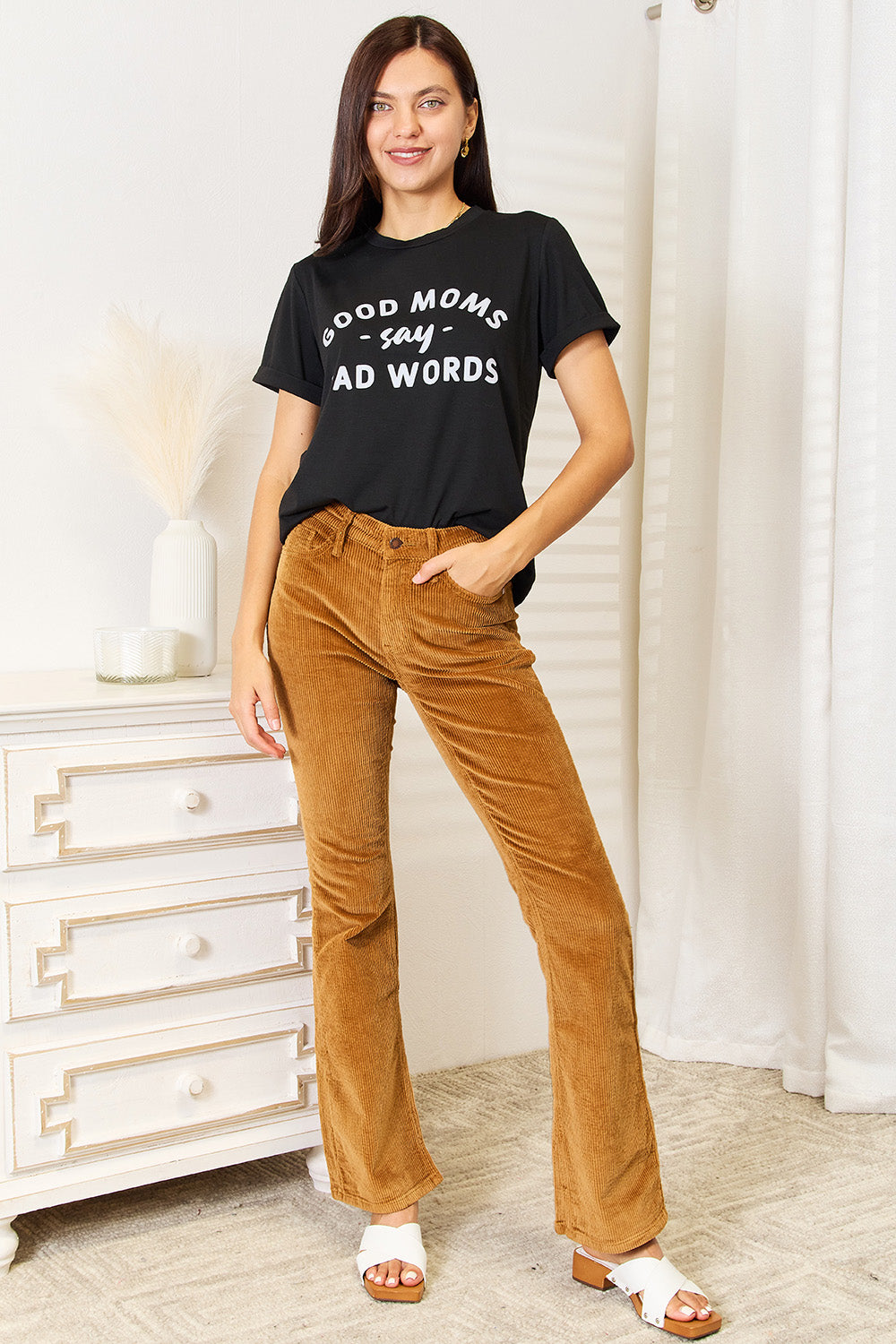 A woman stands in a room with a white wall, where light filters through the blinds, subtly showcasing her rebellious side while wearing the Simply Love GOOD MOMS SAY BAD WORDS Graphic Tee.