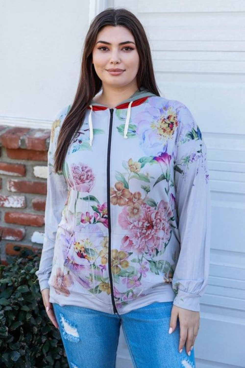 A woman stands outdoors wearing the Blumin Apparel Full Size Floral Zip-Up Hoodie with her hands in the pockets. A leafy green hedge is in the background, adding to the casual chic vibe.