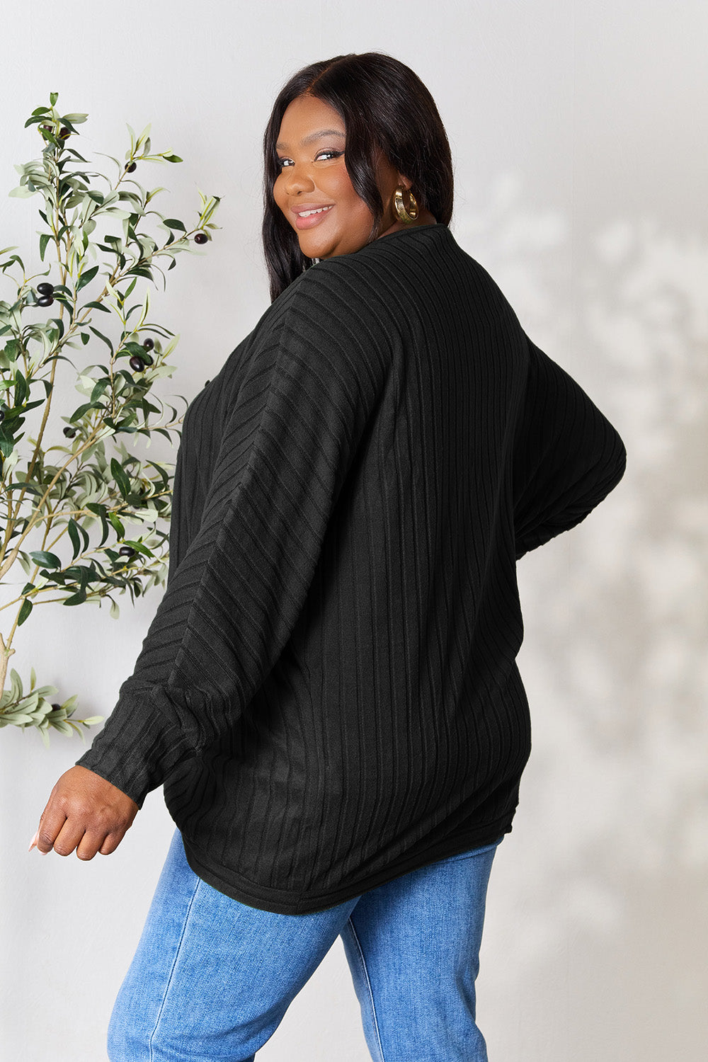 A person with wavy hair, wearing the Basic Bae Full Size Ribbed Cocoon Cardigan over a white top and beige pants, stands in front of a light-colored background with a leafy plant, showcasing an effortlessly basic style.