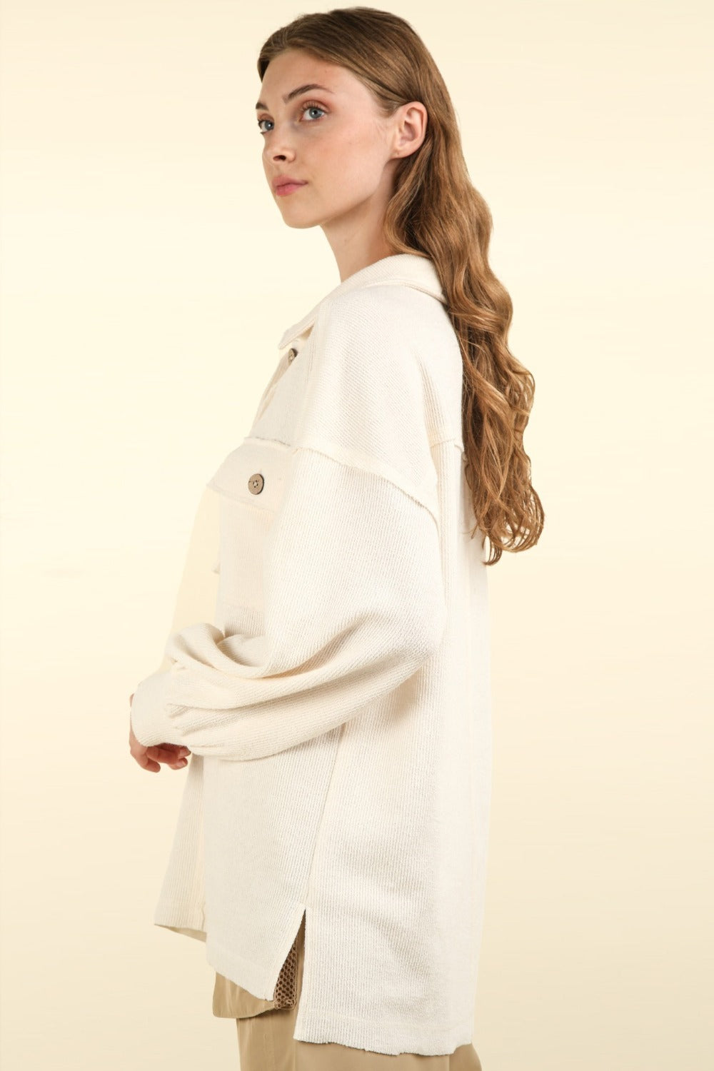 A woman with long hair stands against a neutral background, wearing an oversized VERY J Collared Half Button Knit Top with large pockets in cream.