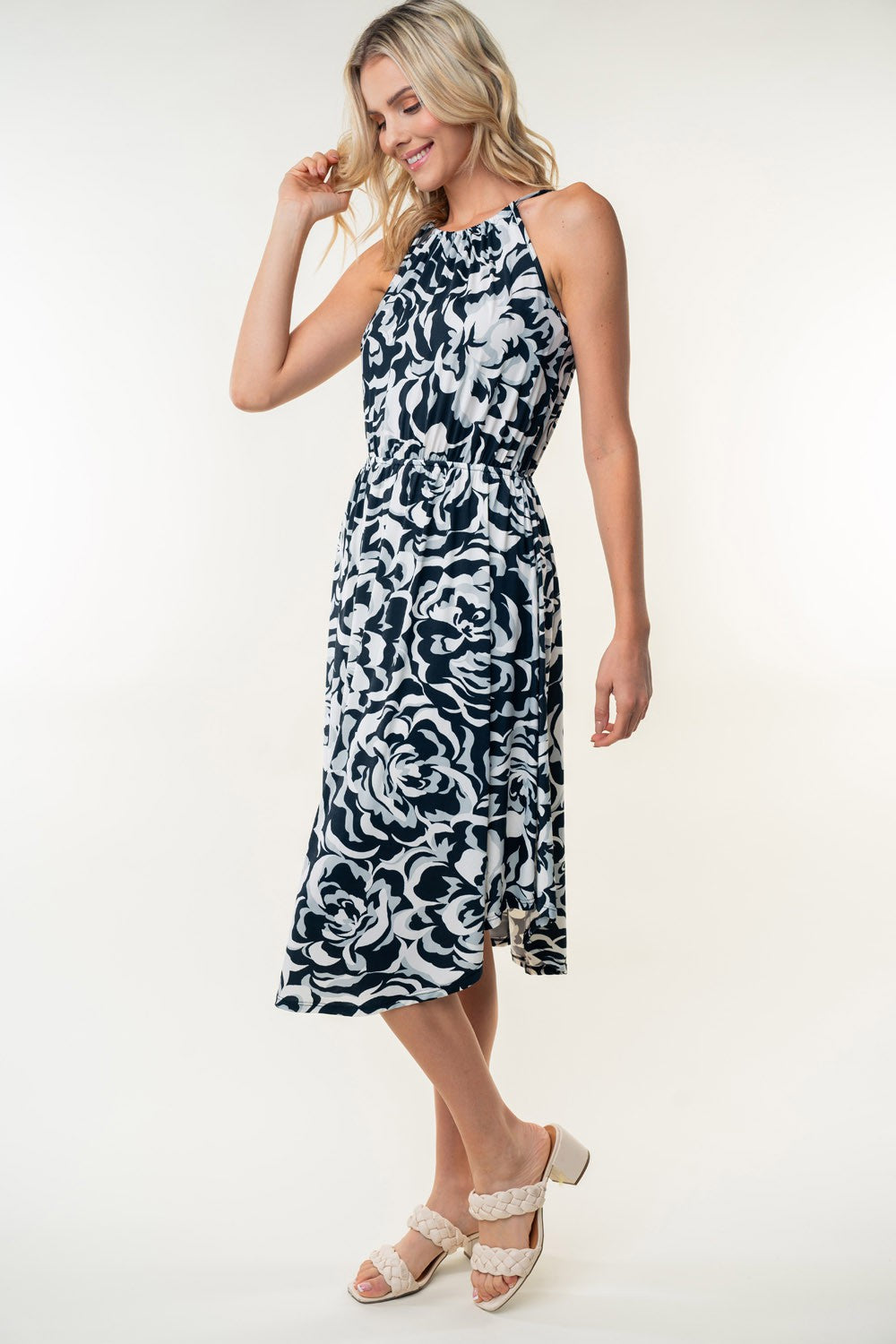 The person wearing a White Birch Tied Ruched Floral Sleeveless Knee Length Dress, featuring a black and white floral pattern, stands and smiles against a neutral background.