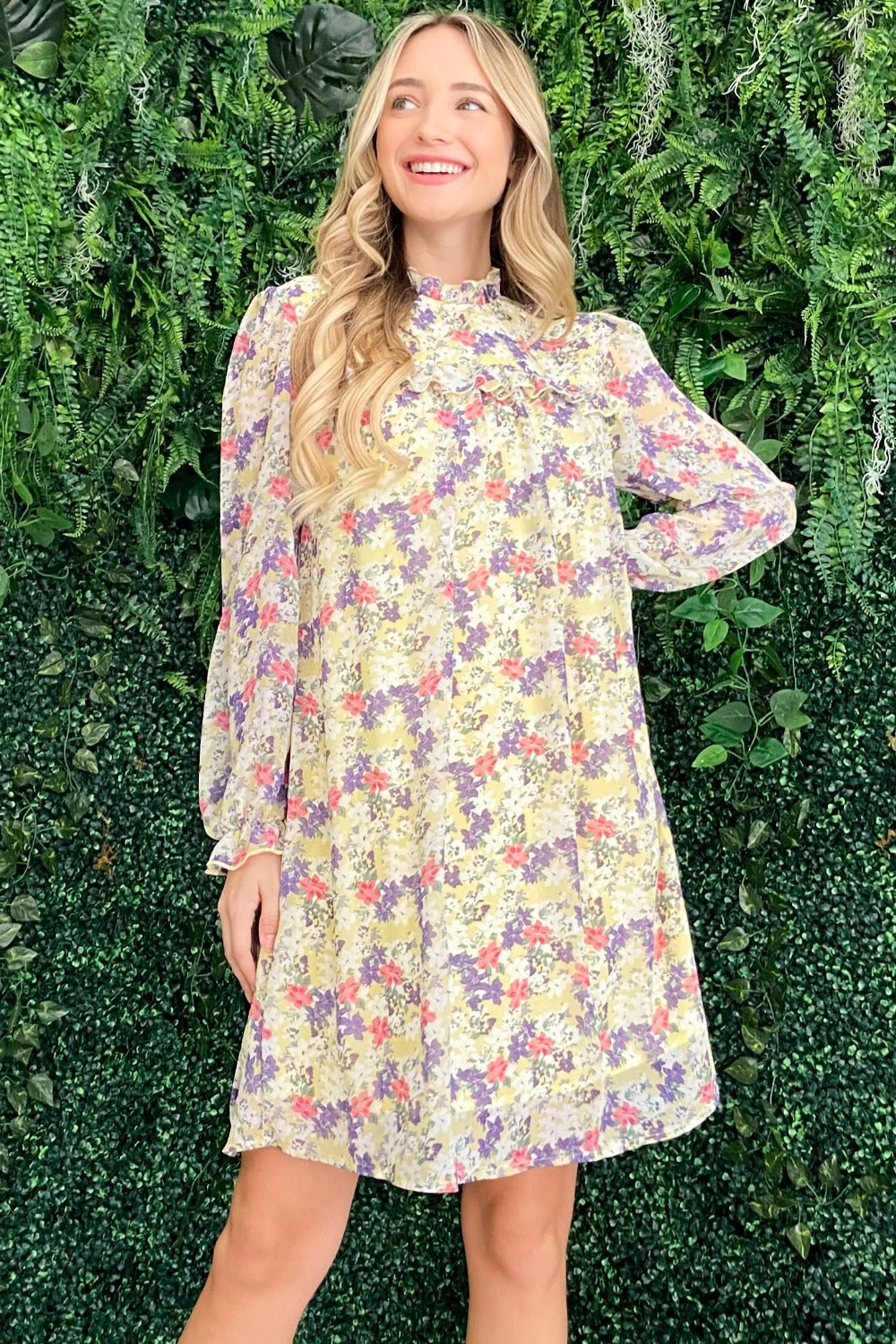 A woman stands in front of a leafy green background wearing the And The Why Floral Mock Neck Flounce Sleeve Dress, smiling and looking to the side.