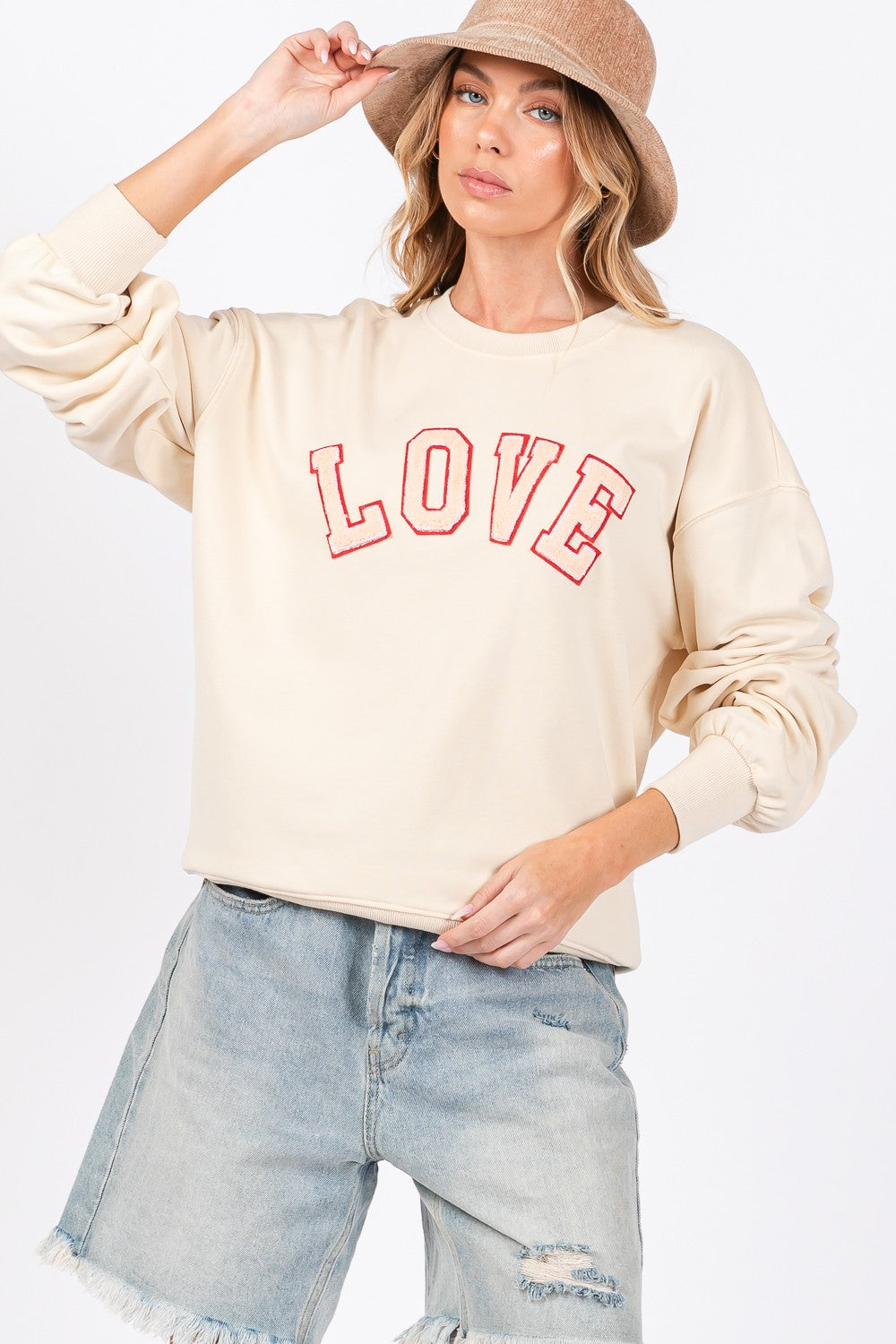 A woman dressed in a SAGE + FIG LOVE Path Applique Drop Shoulder Sweatshirt and a wide-brimmed hat is touching the brim of her hat with both hands, looking to the side and flaunting her cozy and stylish sweatshirt.