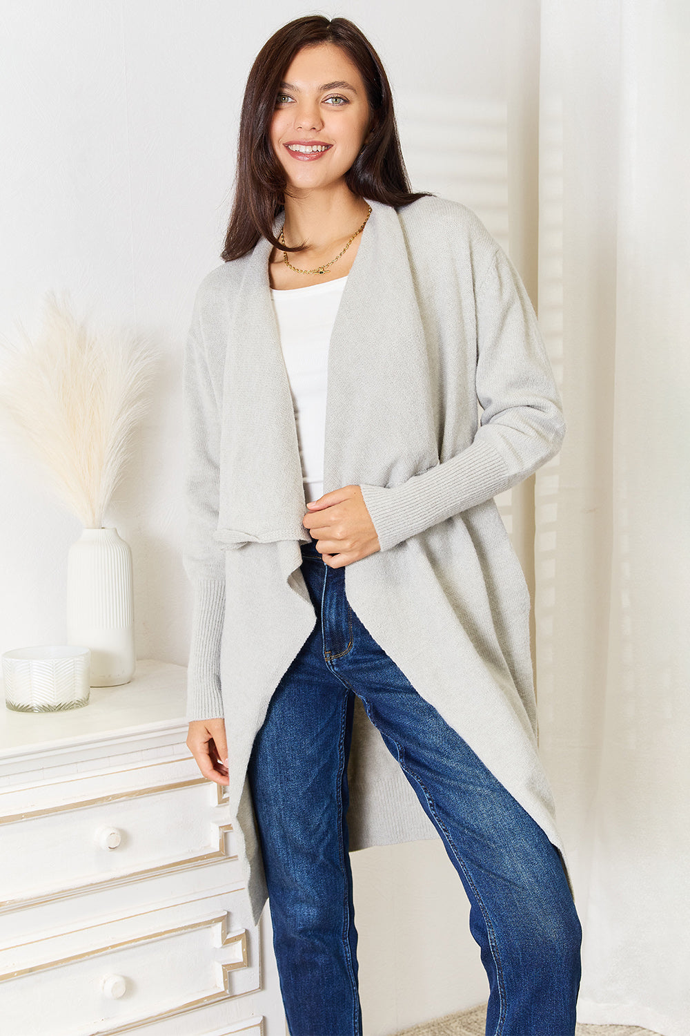 A person with long brown hair wearing a gray Angel Wings Open Front Duster Cardigan with Pockets and blue jeans stands next to a white dresser with a vase and candle on it.