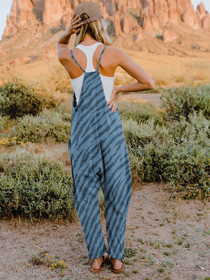 A person wearing a casual white V-neck tank top and a Full Size Printed V-Neck Sleeveless Jumpsuit with a brown animal print zebra-stripe pattern stands outdoors with one hand on their hat and the other in their pocket, set against a rocky landscape backdrop.