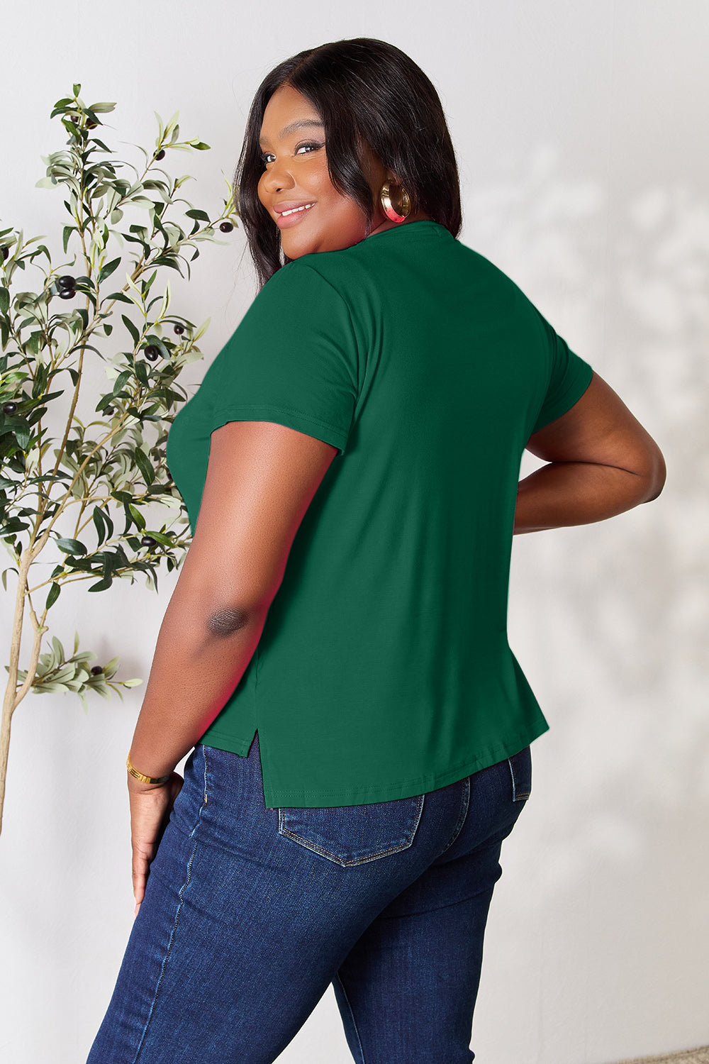 A woman with long hair, smiling with her right hand in her pocket and standing next to a plant, is wearing the Basic Bae Full Size Round Neck Short Sleeve T-Shirt in black along with beige pants made of rayon spandex.