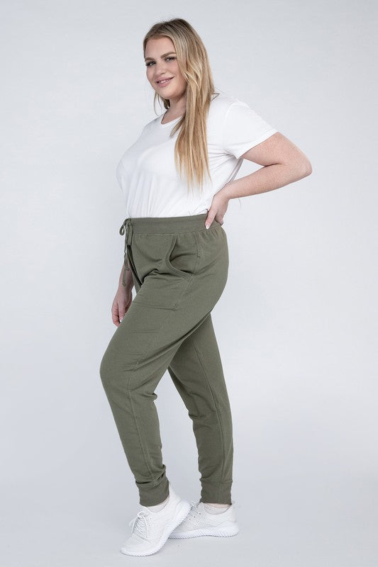 A person stands against a white background, wearing a white t-shirt, grey Plus-Size Jogger Pants with an adjustable waistband, and white sneakers. They have long blonde hair and are posing with one hand on their hip.