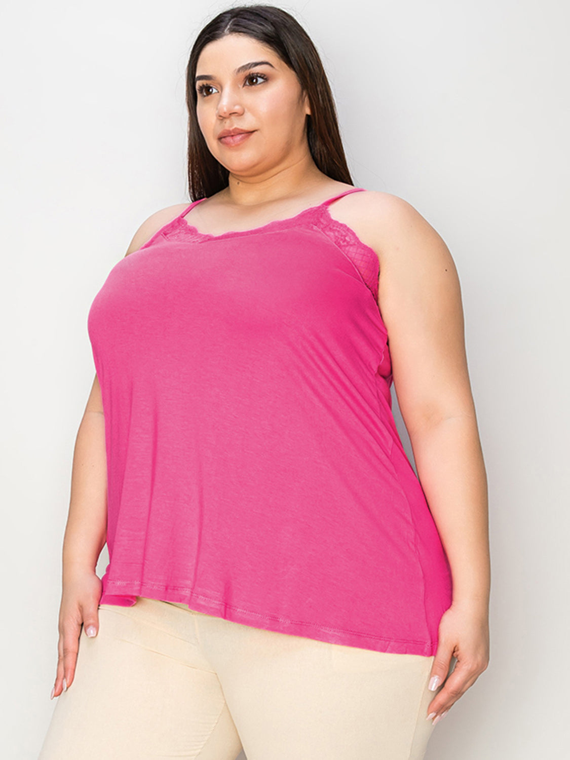 A woman with long hair wearing sunglasses, a sleeveless orange Basic Bae Full Size Lace Trim V-Neck Cami, and blue jeans stands against a concrete wall. She is looking to the side and holding one arm up, touching her sunglasses. Her outfit, available in plus sizes and made from a comfortable rayon blend, perfectly complements her effortless style.