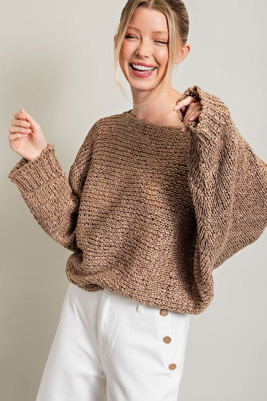 A woman smiles while wearing the Loose Fit Knit Top featuring a boat neckline, paired with white pants with button embellishments on the side. She stands against a plain background.