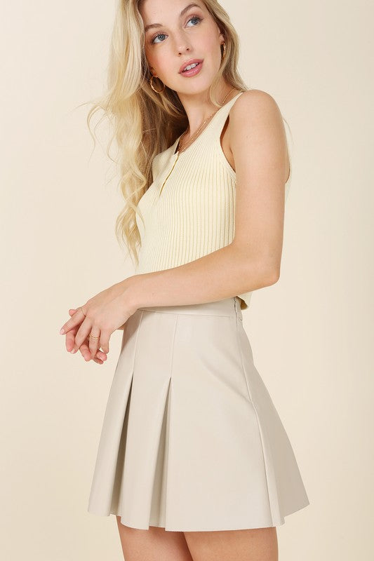 A woman in a beige tank top stands against a light background, looking to her right while wearing the Vegan Leather Pleated Mini Skirt featuring a side zipper.