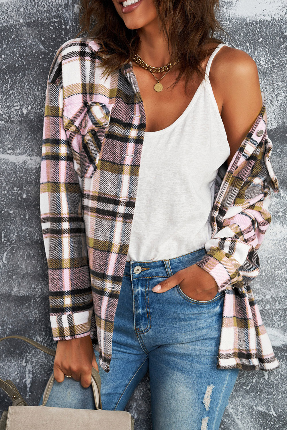 A person wearing a Brown Plaid Button Up Long Sleeve Flannel Shacket layered over a white top with ripped jeans and holding a brown handbag stands against a textured gray wall.