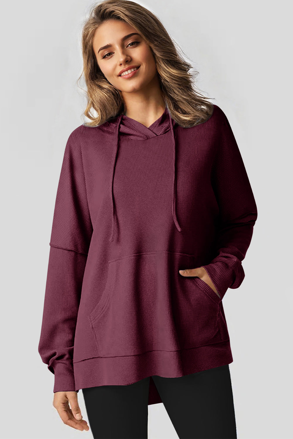 A woman in a Red Dahlia Waffle Knit Fleece Lined High Low Oversized Hoodie and black shorts is seen from behind, standing against a plain gray background.