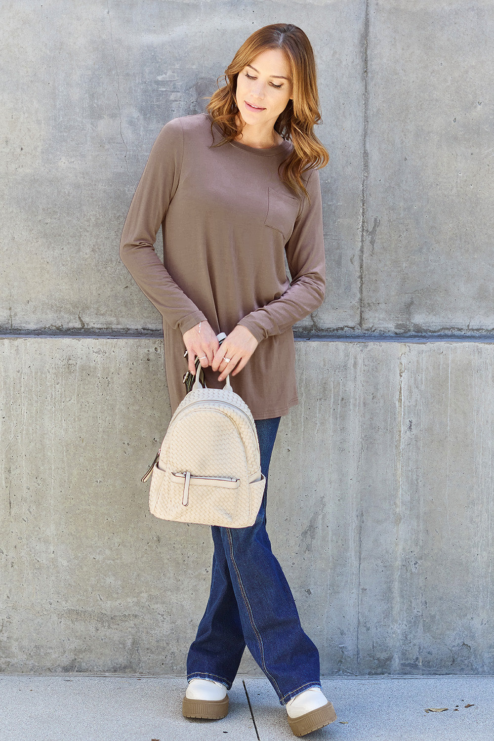 A woman with long hair wearing sunglasses, a Basic Bae Full Size Round Neck Long Sleeve Top in turquoise, and jeans is standing in front of a concrete wall with one hand on her hip.