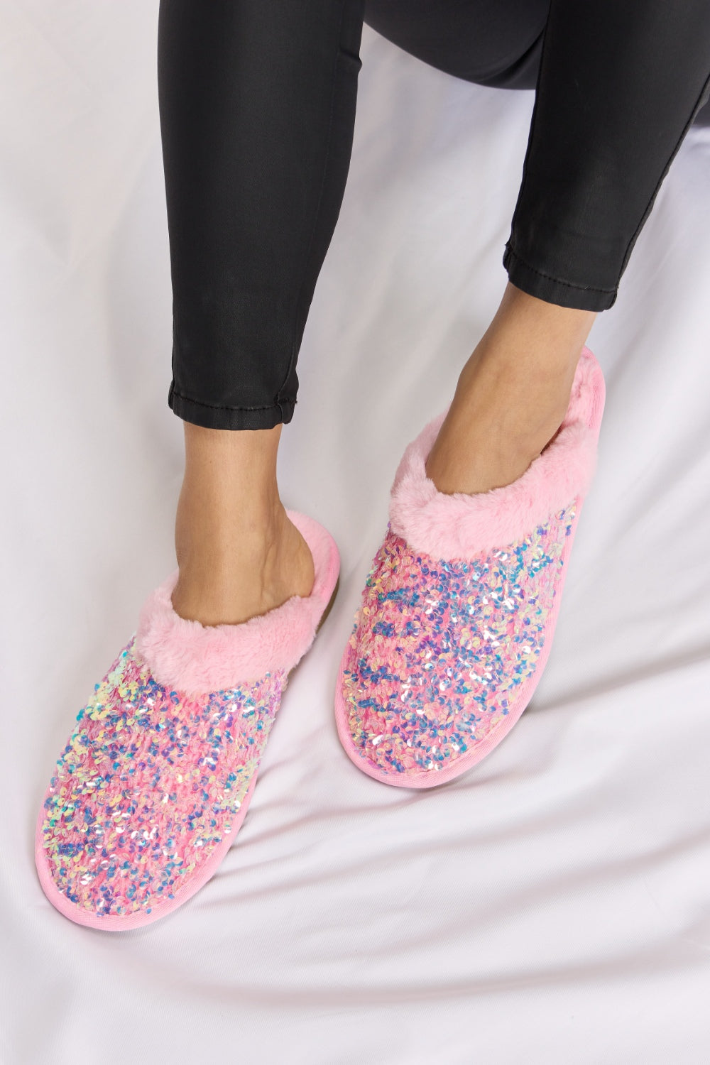 Person wearing cozy and luxurious Forever Link Sequin Plush Round Toe Slippers with fuzzy trim and black pants, sitting on a white surface.