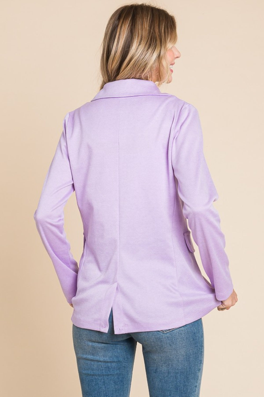 A person with long, wavy hair is wearing the Culture Code Button Up Long Sleeve Slit Blazer in light purple over a white top and blue jeans, standing against a beige background.