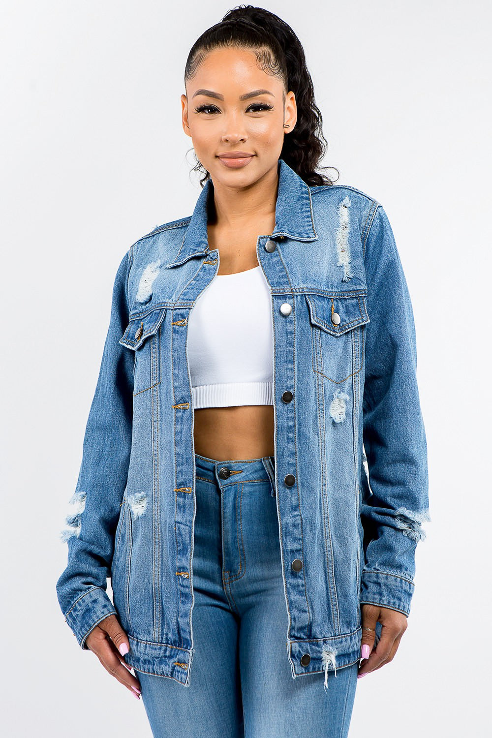 A woman in an American Bazi Full Size Button Up Distressed Denim Jacket with a vintage look stands against a plain background, pairing it perfectly with a white crop top and denim jeans.