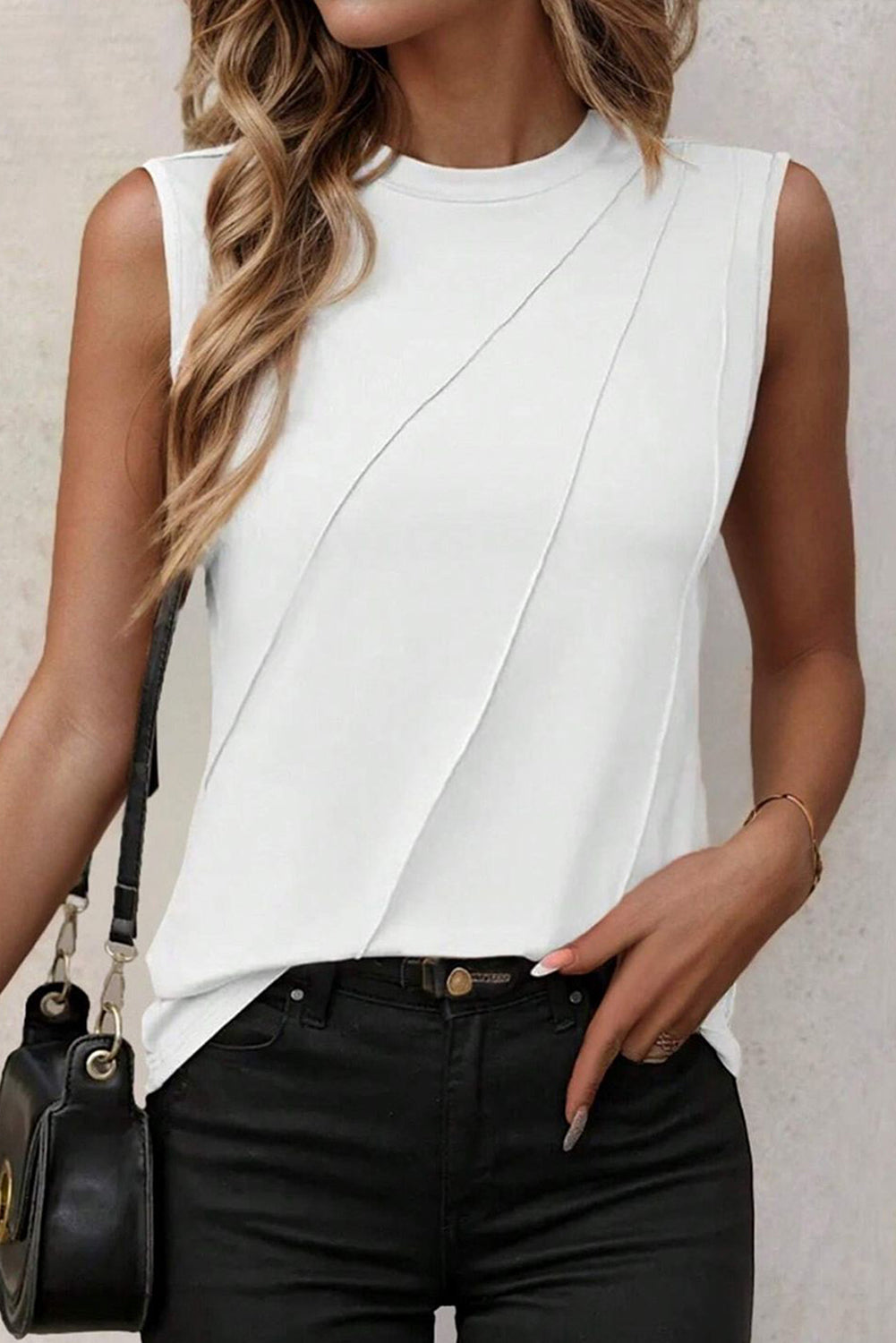 A woman is wearing a White Solid Color Crew Neck Pleated Tank Top made from breathable fabric with a diagonal seam design, black jeans, and a black crossbody bag. Her left hand rests on her waist.