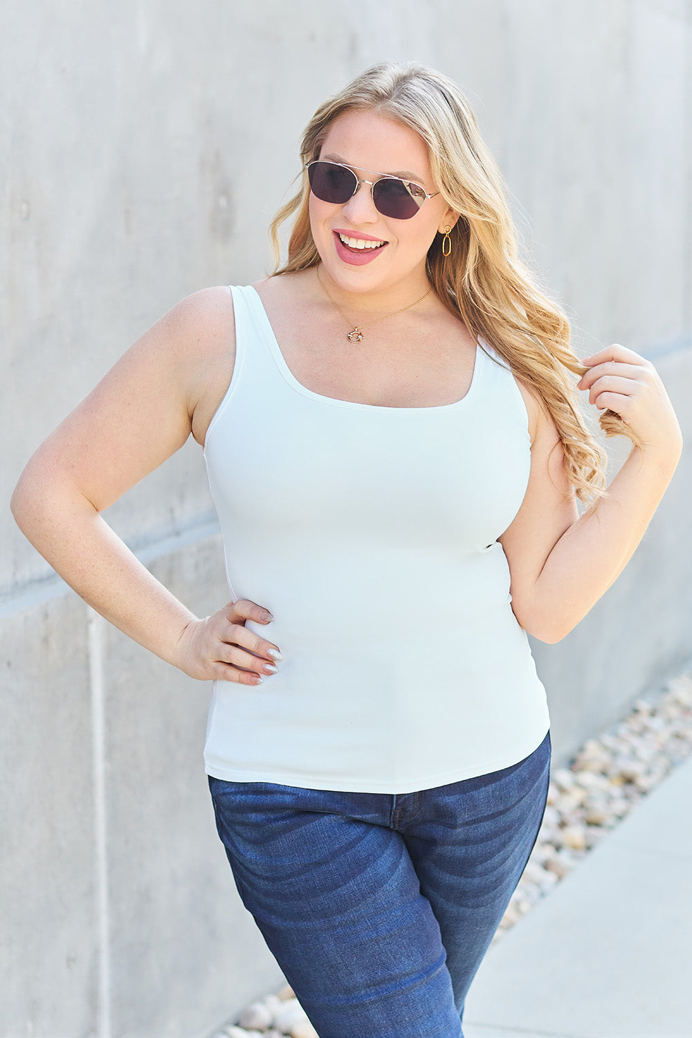 A woman with brown hair, wearing sunglasses, blue jeans, and the Basic Bae Full Size Square Neck Wide Strap Tank in black stands against a concrete wall, showcasing a basic style.