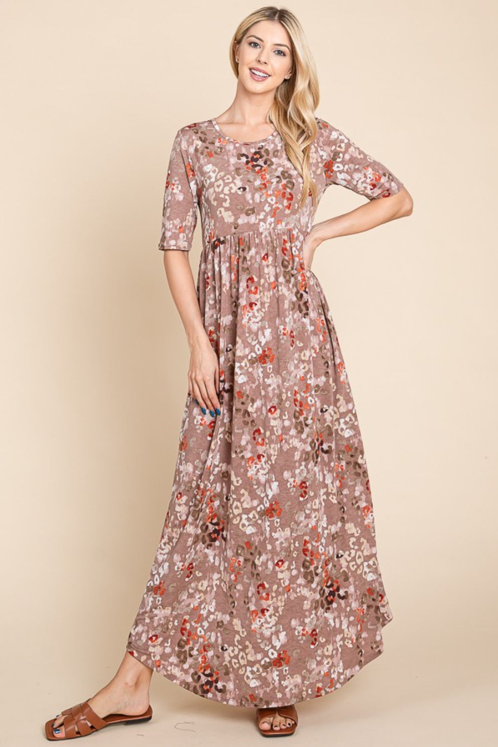 A woman stands against a beige background wearing the light brown, floral BOMBOM Printed Shirred Maxi Dress and brown sandals. She has long blond hair and is looking to her right with one hand slightly raised. The versatile dress offers a flattering fit perfect for any occasion.