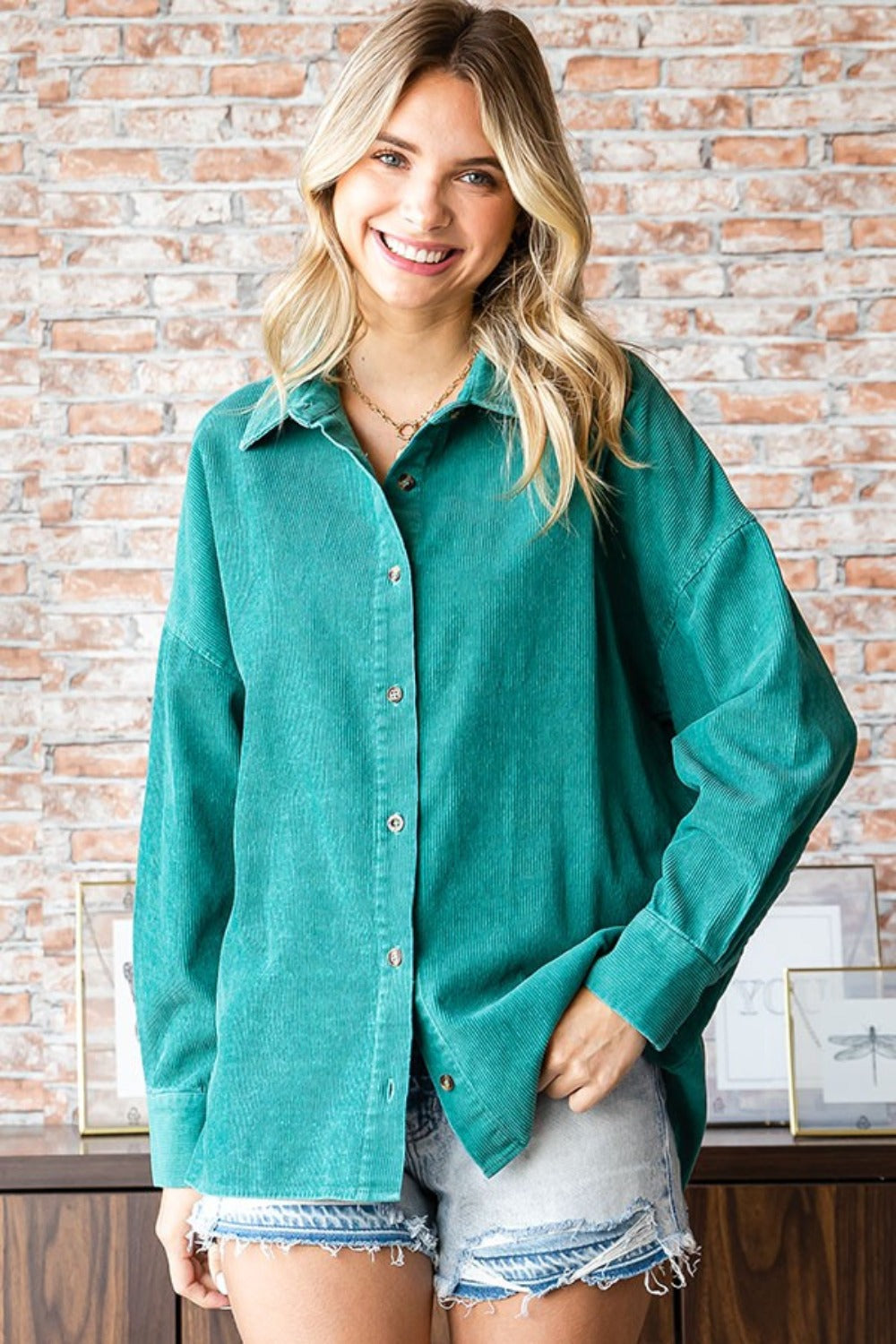 A woman wearing the First Love Washed Corduroy Button Down Long Sleeve Shirt in green, a white top, and denim shorts poses indoors. She stands in front of a brick wall with a leafy plant on the side.