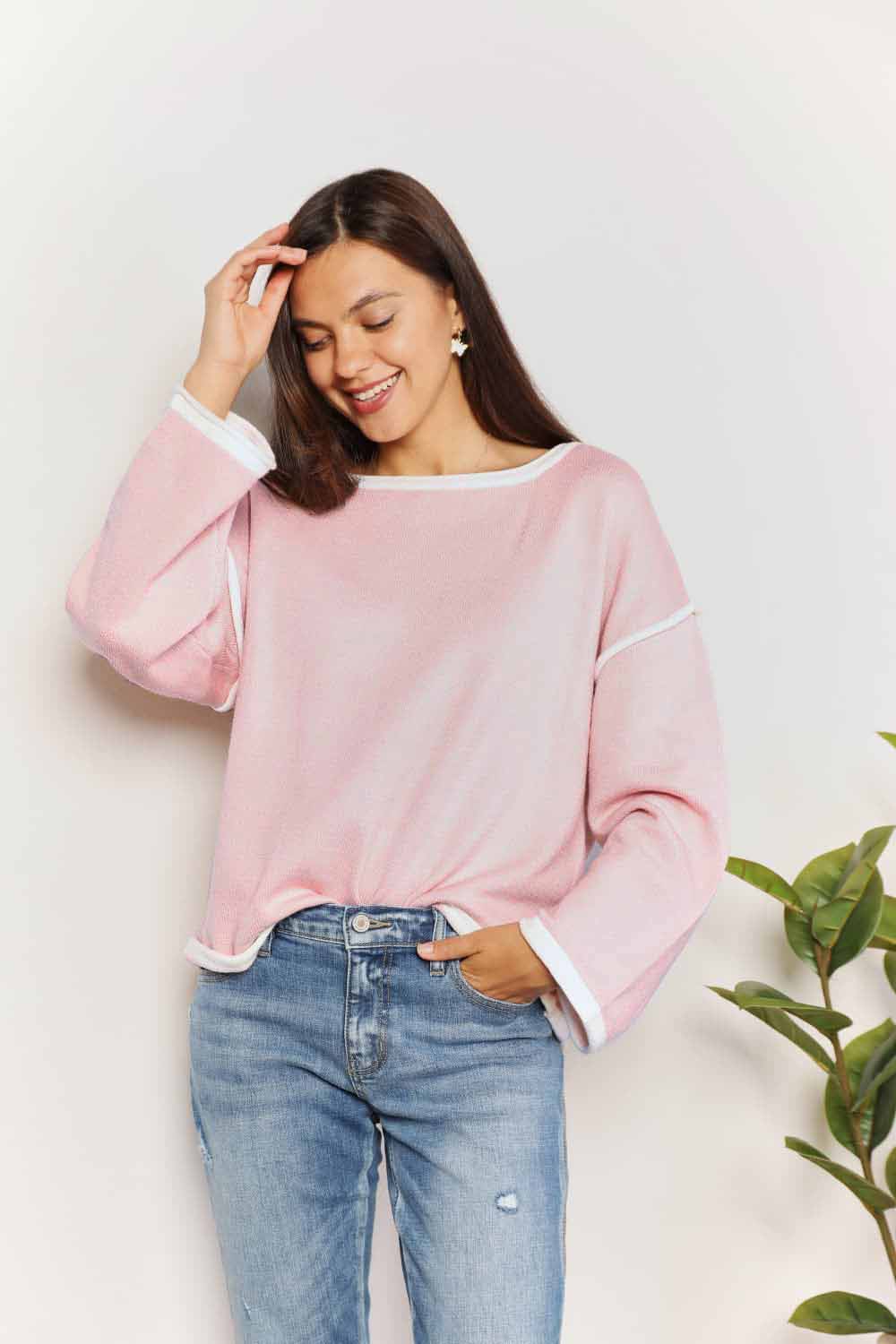 A woman with long curly hair is wearing the Angel Wings Contrast Detail Dropped Shoulder Knit Top in pink, paired with blue jeans, standing against a light background.