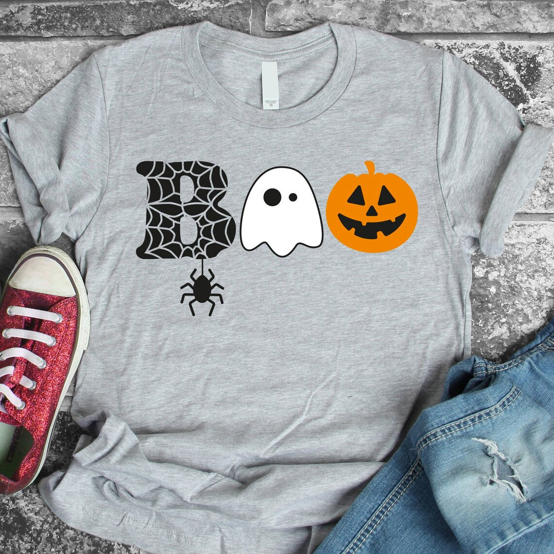 Introducing our Boo Halloween T-shirt: a Gray Bella Canvas tee adorned with the word "Boo," featuring a spider web "B", a ghost "O", and a pumpkin "O". Laid on a stone surface, this shirt is pictured next to red sneakers and blue jeans, highlighting its soft textile flex print.