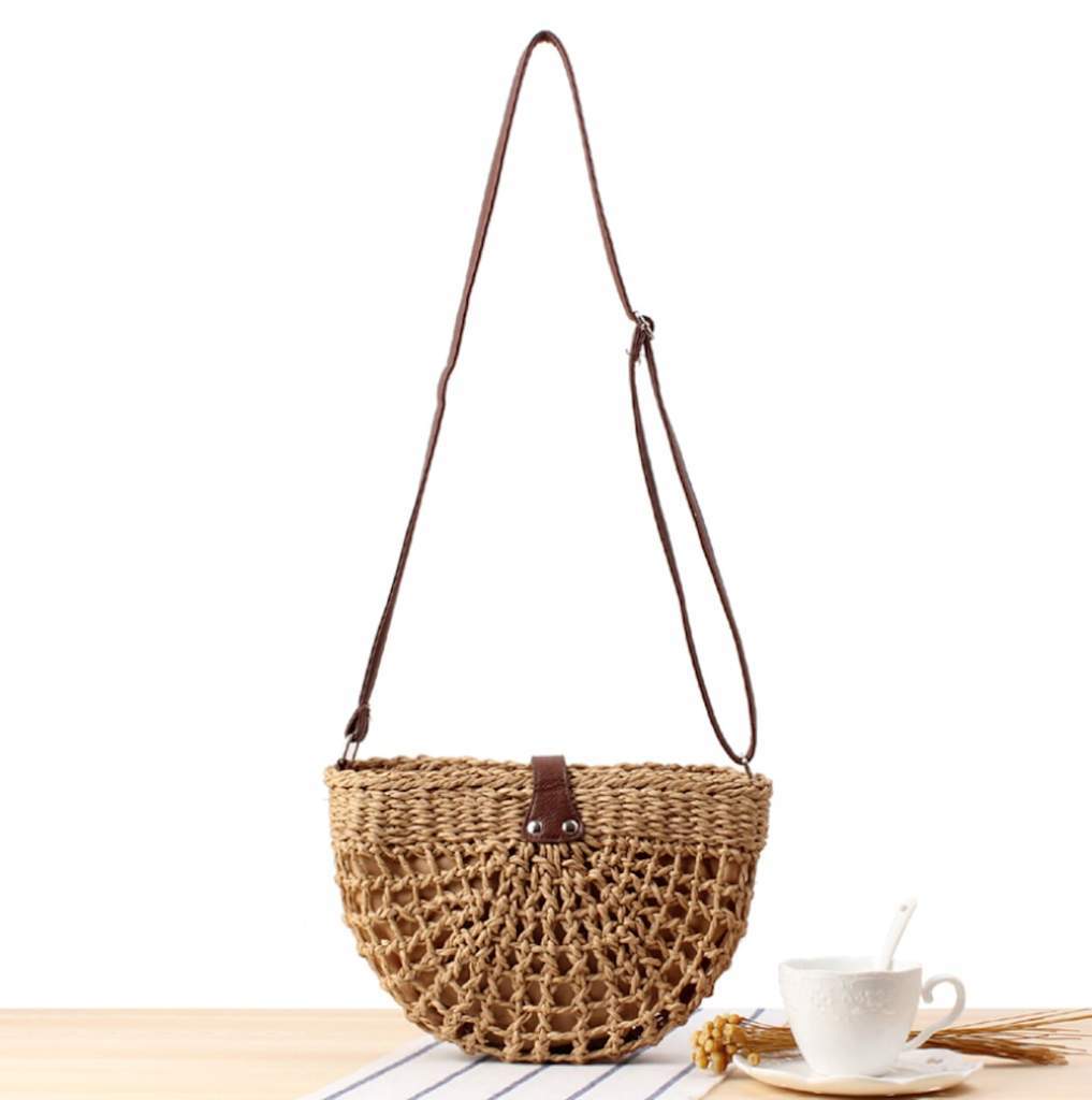 Half Moon Straw Crossbody Bag with a vegan leather strap and clasp, displayed on a table beside a cup and saucer.