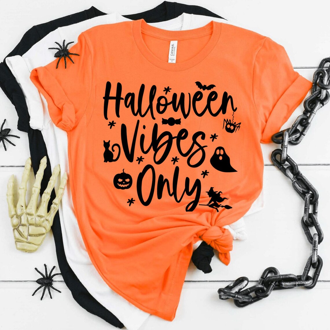 The Halloween Vibes Only Halloween T-shirt is displayed on a white surface, featuring an orange color with the text "Halloween Vibes Only" surrounded by Halloween-themed graphics, including a skeleton hand, black spiders, and black chains. This premium quality t-shirt boasts a soft textile flex print for a comfortable retail fit.