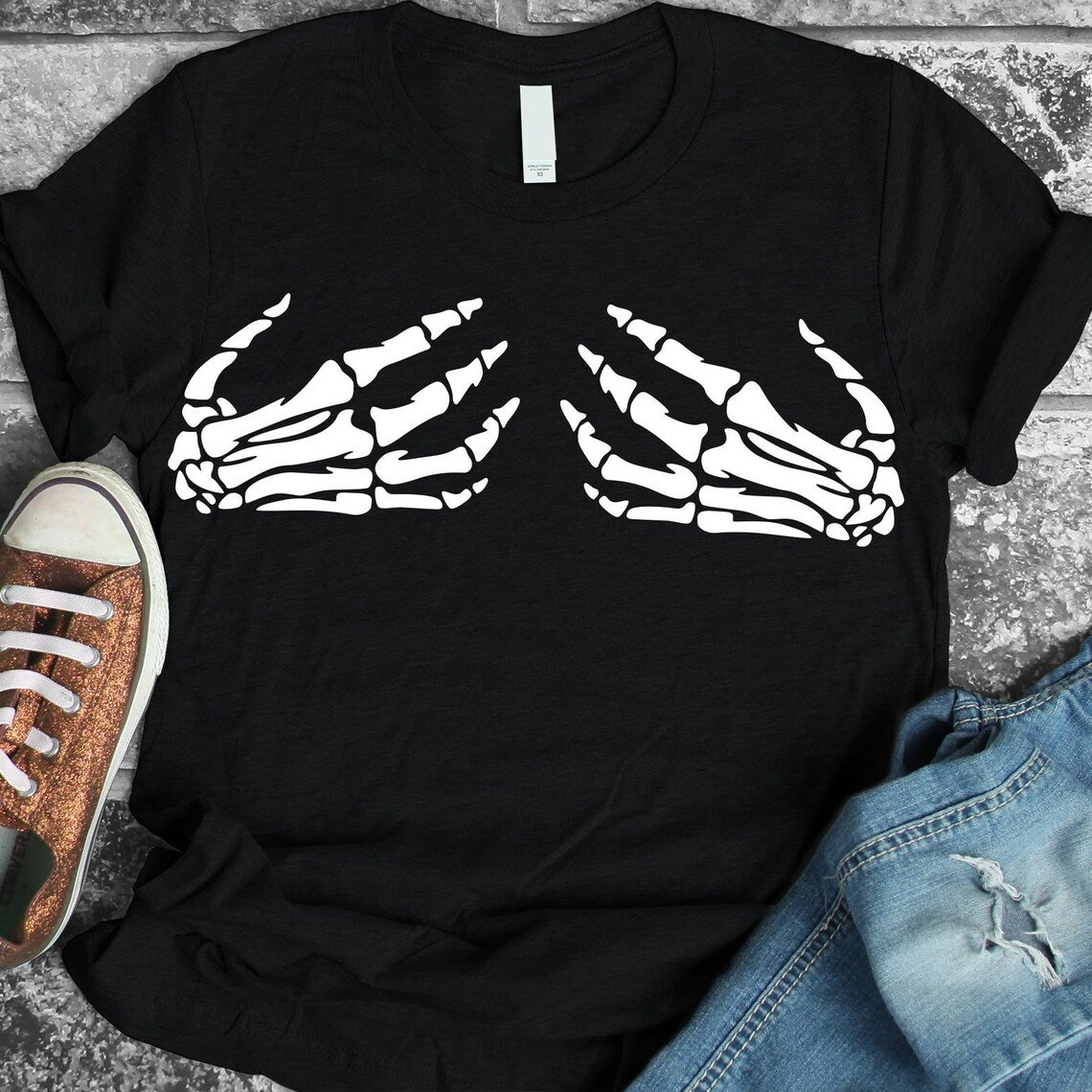 Skeleton Hand Halloween T-shirt, featuring premium quality black fabric with a soft textile flex print of white skeleton hand graphics on the chest, displayed on a stone surface next to a pair of brown shoes and blue jeans.