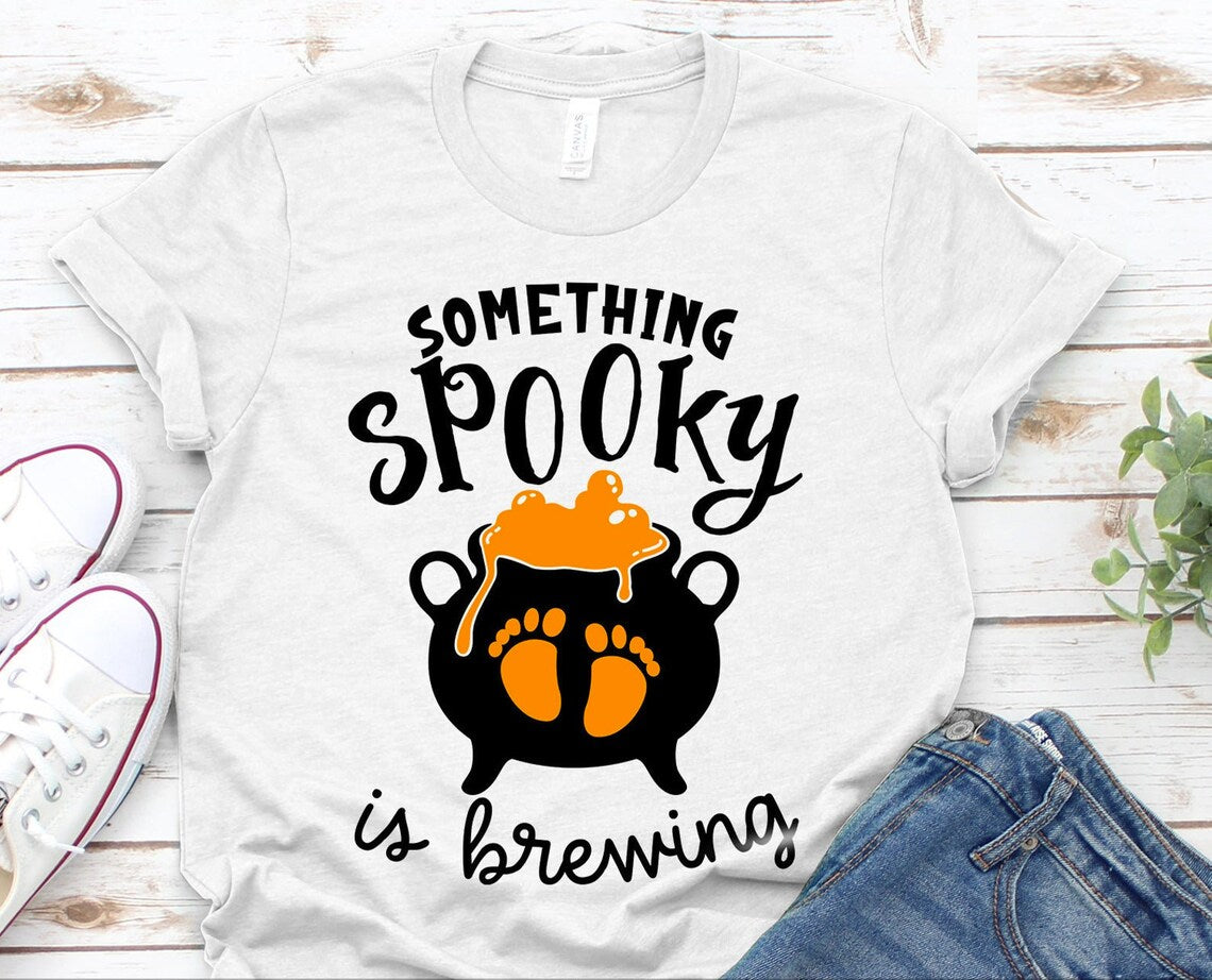 The "Something Spooky Is Brewing Halloween T-shirt" features the text "Something Spooky is Brewing" along with a graphic of a bubbling cauldron. Crafted from soft textile flex print for lasting comfort, this premium quality white t-shirt is showcased on a wooden surface alongside a pair of sneakers and denim jeans.