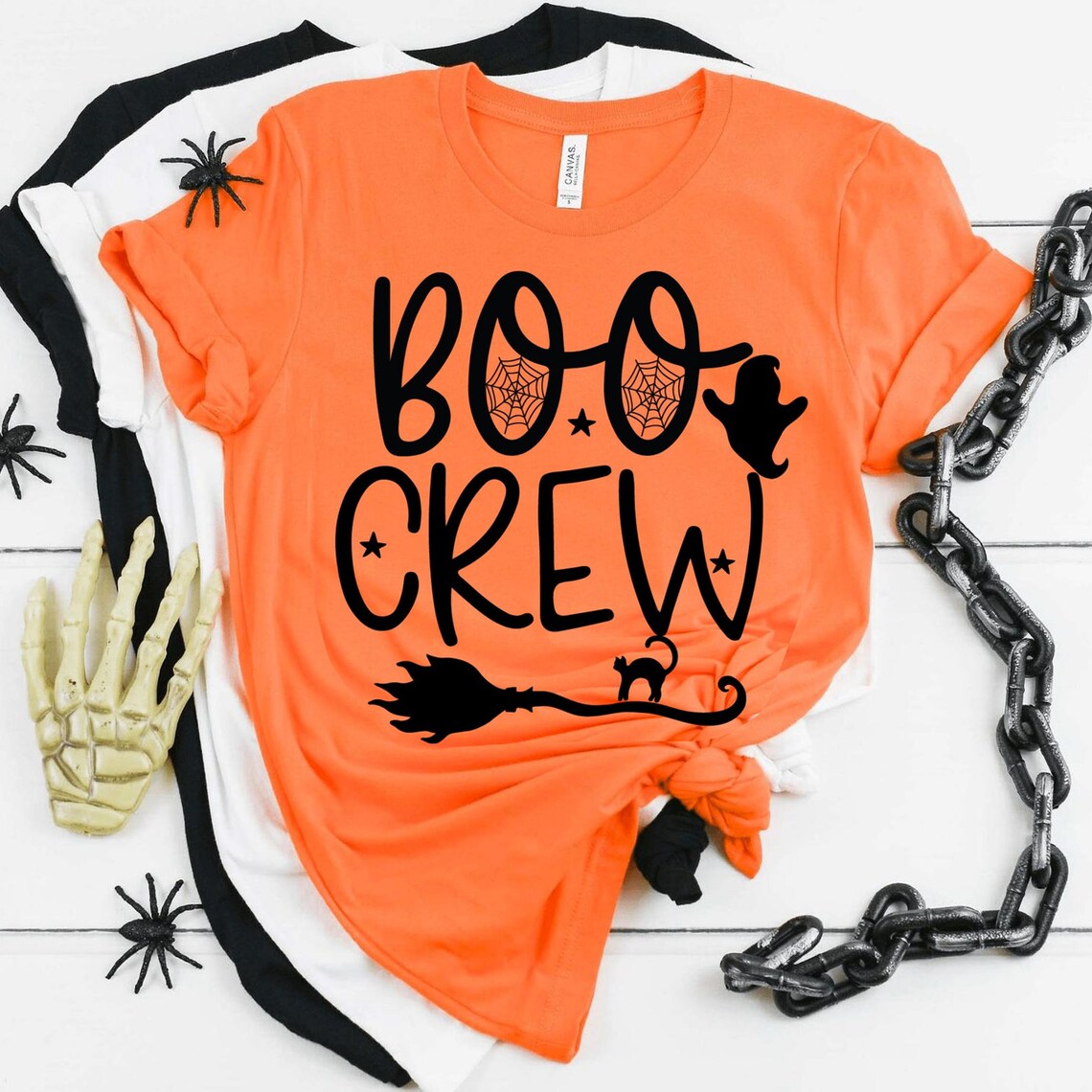 Three Boo Crew T-shirts with Halloween designs feature text "Boo Crew," along with illustrations of a ghost, spider webs, and a witch's broom. Props include plastic spiders, a skeletal hand, and a black chain on a white background. Each shirt is crafted from soft textile flex print to ensure premium quality comfort.