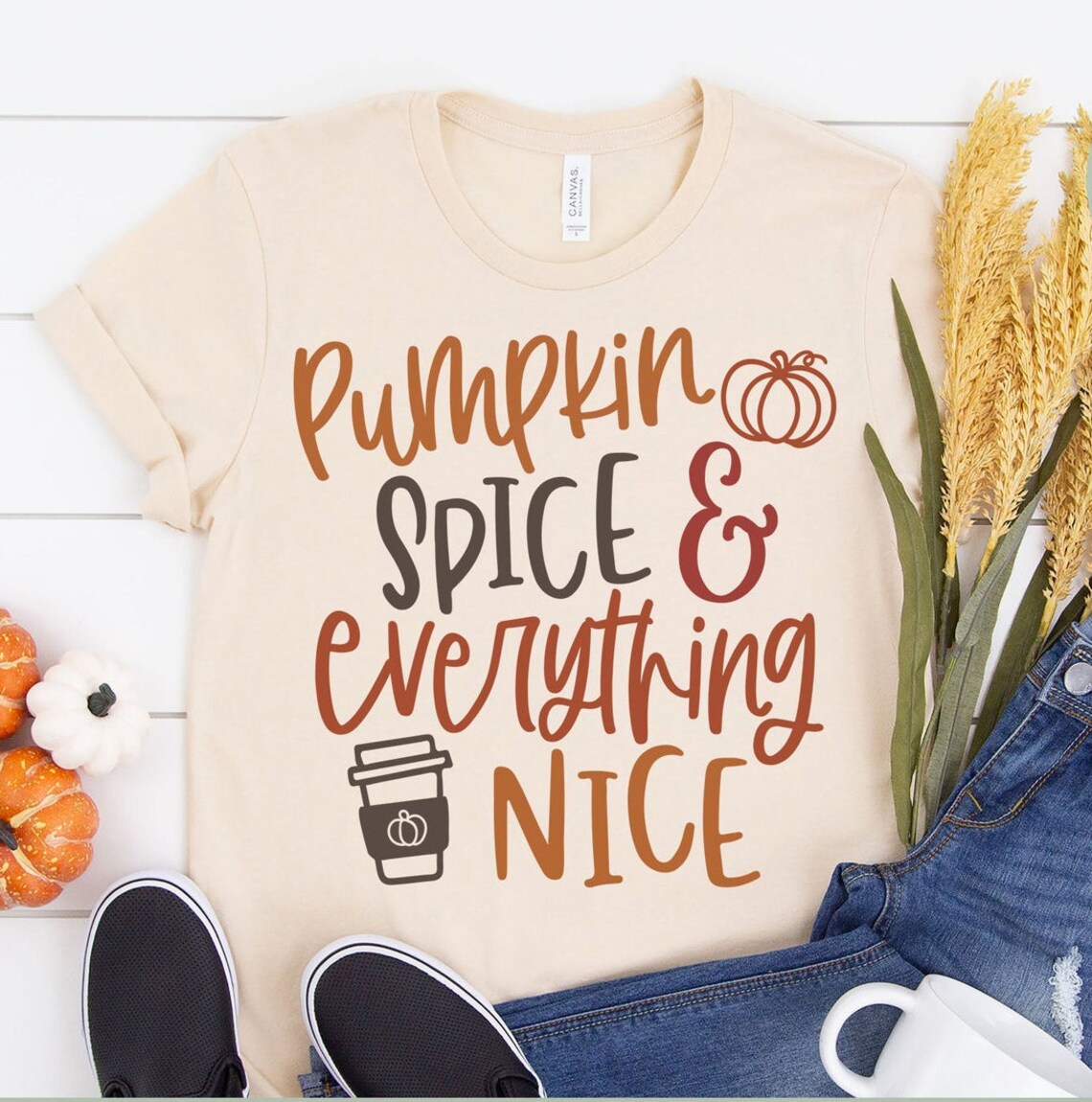 Get ready for autumn with the Pumpkin Spice Everything Nice Halloween T-shirt, a premium quality beige top featuring "Pumpkin Spice & Everything Nice" text surrounded by charming autumn decorations. Pair it perfectly with blue jeans, black shoes, and a white cup for that ultimate seasonal look.