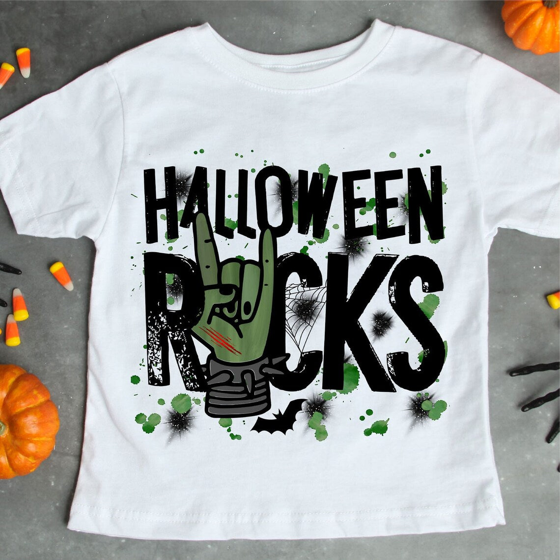 The Halloween Rocks Halloween T-shirt is a premium quality Bella Canvas white t-shirt featuring the text "HALLOWEEN ROCKS" in black. It showcases a green hand making a rock sign, surrounded by Halloween-themed graphics such as a spider web, bats, and black splatters. The shirt features a soft textile flex print for ultimate comfort.