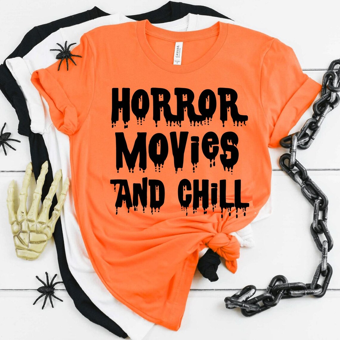 Stacked orange and white "Horror Movies And Chill" t-shirts by Bella Canvas, with the stylish text displayed on the orange shirt. The scene is accented with plastic spiders, a skeleton hand, and chains placed around them. These premium quality t-shirts offer ultimate comfort with their soft textile flex print.