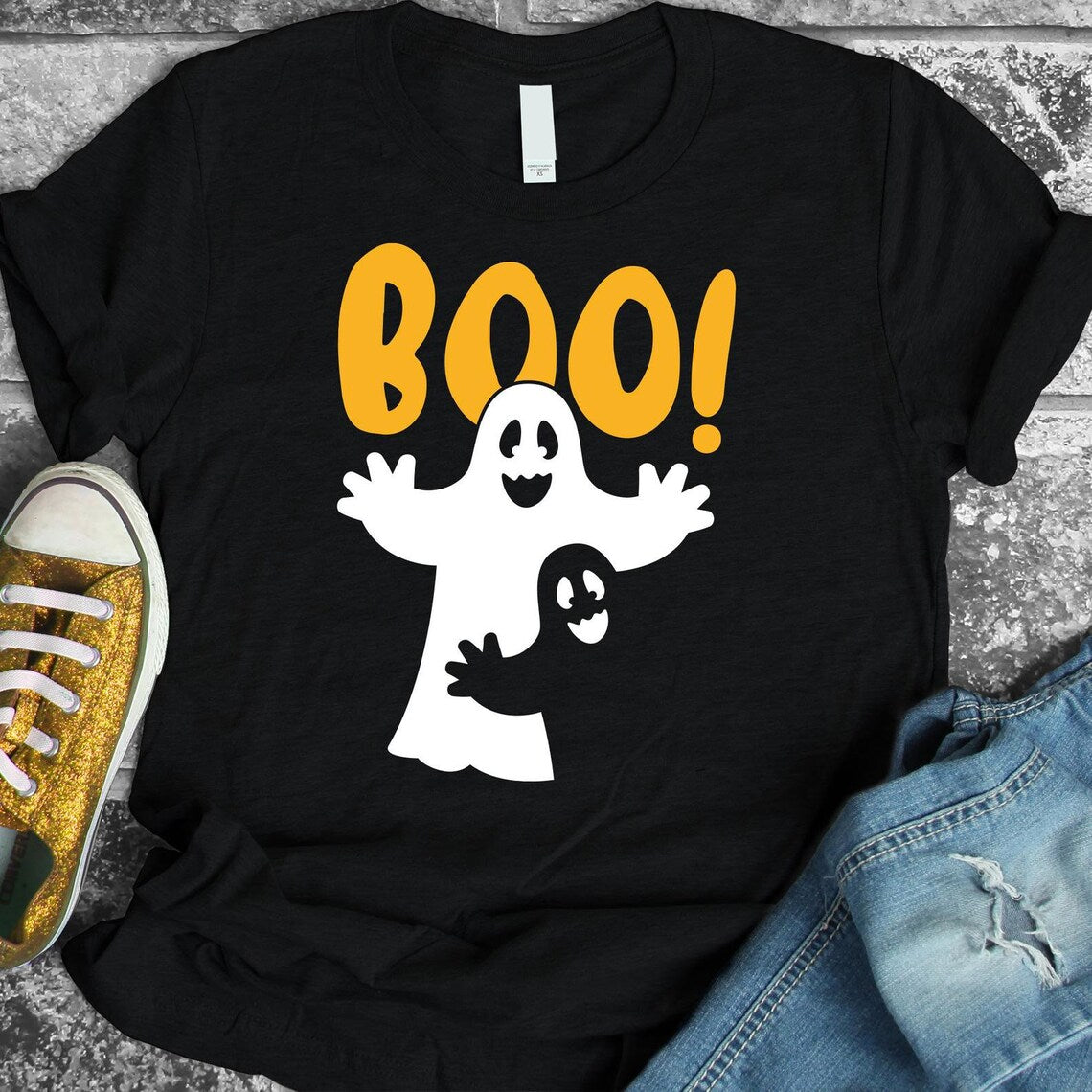 The Boo Halloween T-shirt by Bella Canvas features a graphic of two white cartoon ghosts and the word "BOO!" in orange text. The soft cotton shirt, displayed on a stone surface alongside a yellow sneaker and blue jeans, highlights its premium quality.