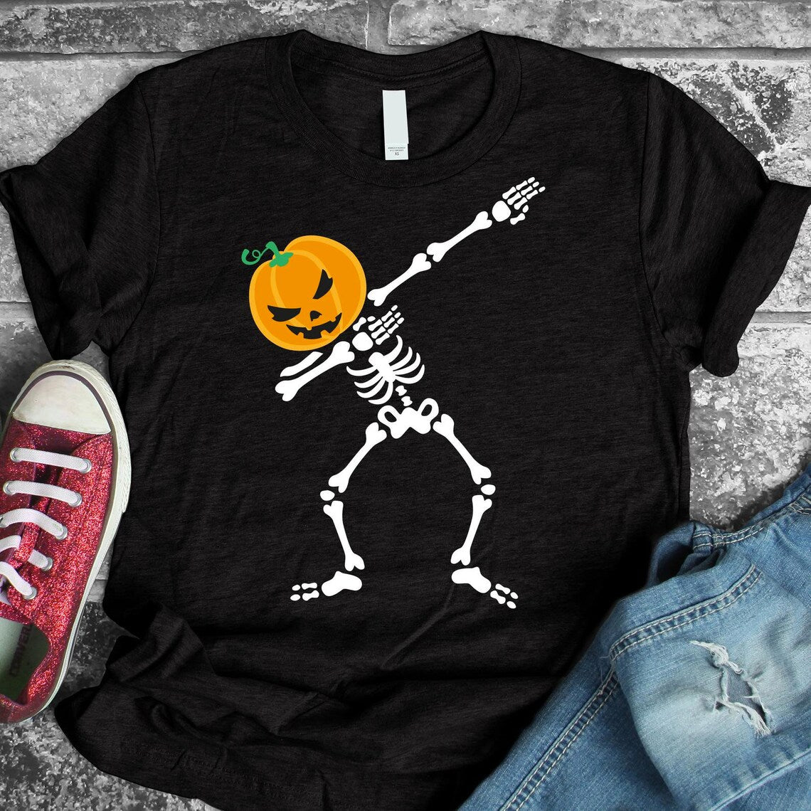 The Skeleton Pumpkin Dabbing Halloween T-shirt is a premium quality black cotton tee from Bella Canvas, showcasing a cartoon skeleton with a pumpkin head dabbing. Pictured alongside blue jeans and a single red sneaker on a gray textured background, it features a soft textile flex print for unmatched comfort.