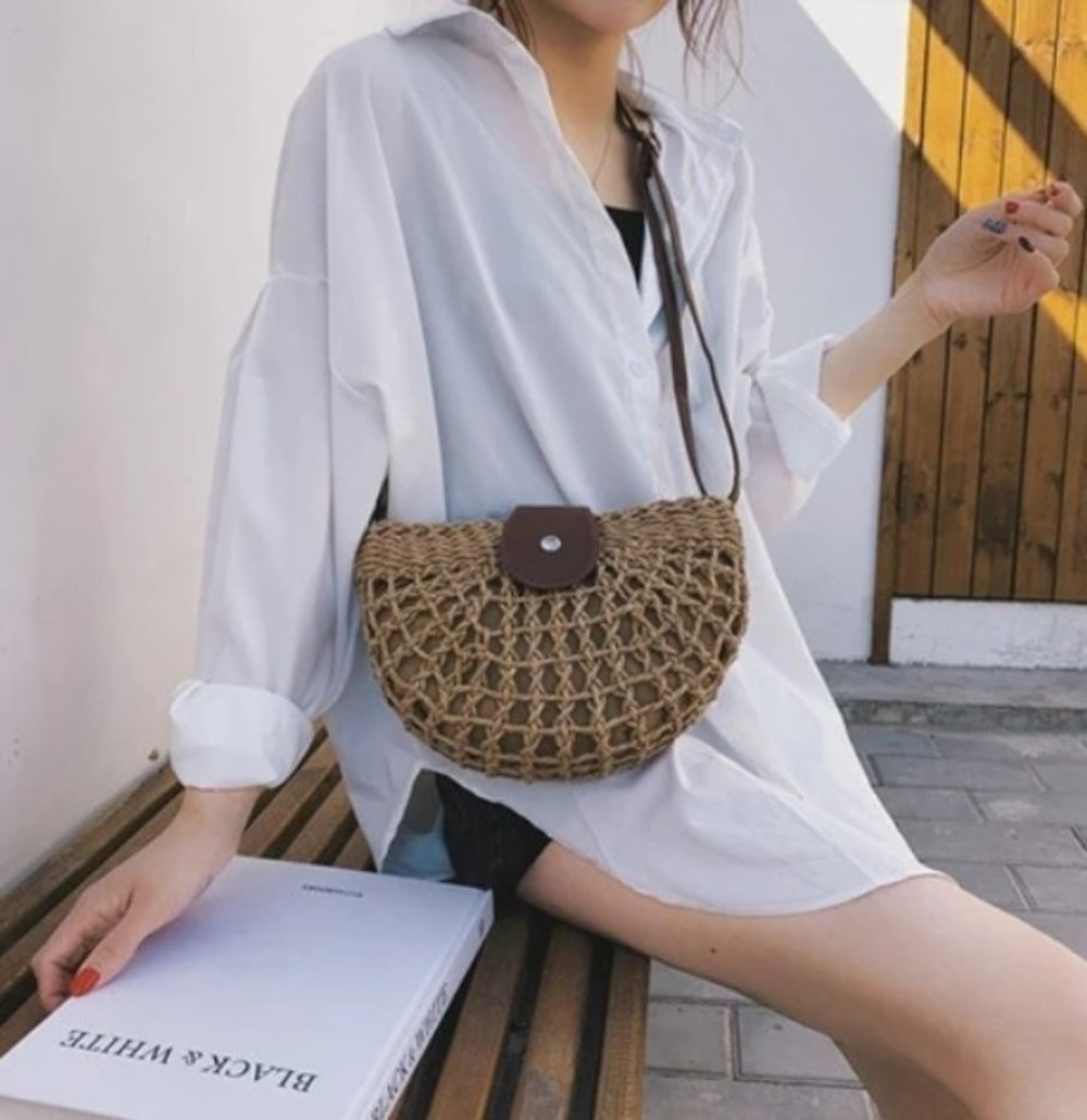 Half Moon Straw Crossbody Bag with a vegan leather strap and clasp, displayed on a table beside a cup and saucer.