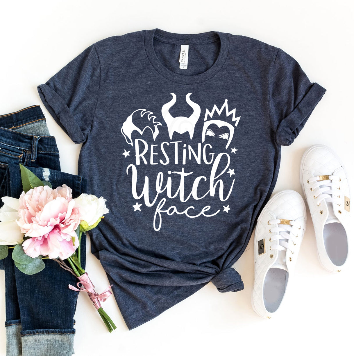 The Resting Witch Face T-shirt in dark gray, featuring the phrase "Resting Witch Face" surrounded by witch-themed graphics, is showcased with a pair of blue jeans, white shoes, and a bouquet of flowers. This premium quality tee provides a retail fit and soft feel, making it ideal for casual outings.