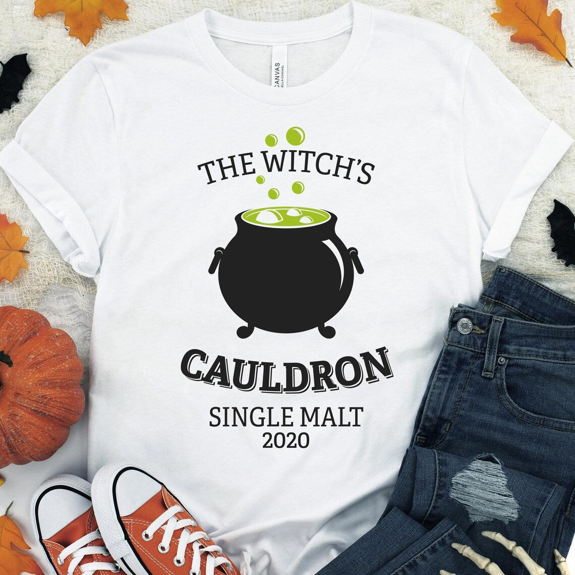 The Witch Halloween T-shirt, made from premium quality cotton and featuring a soft textile flex print of a bubbling cauldron along with the text "The Witch's Cauldron Single Malt 2020," is showcased alongside ripped jeans and orange sneakers. Autumn leaves and pumpkins are scattered around.