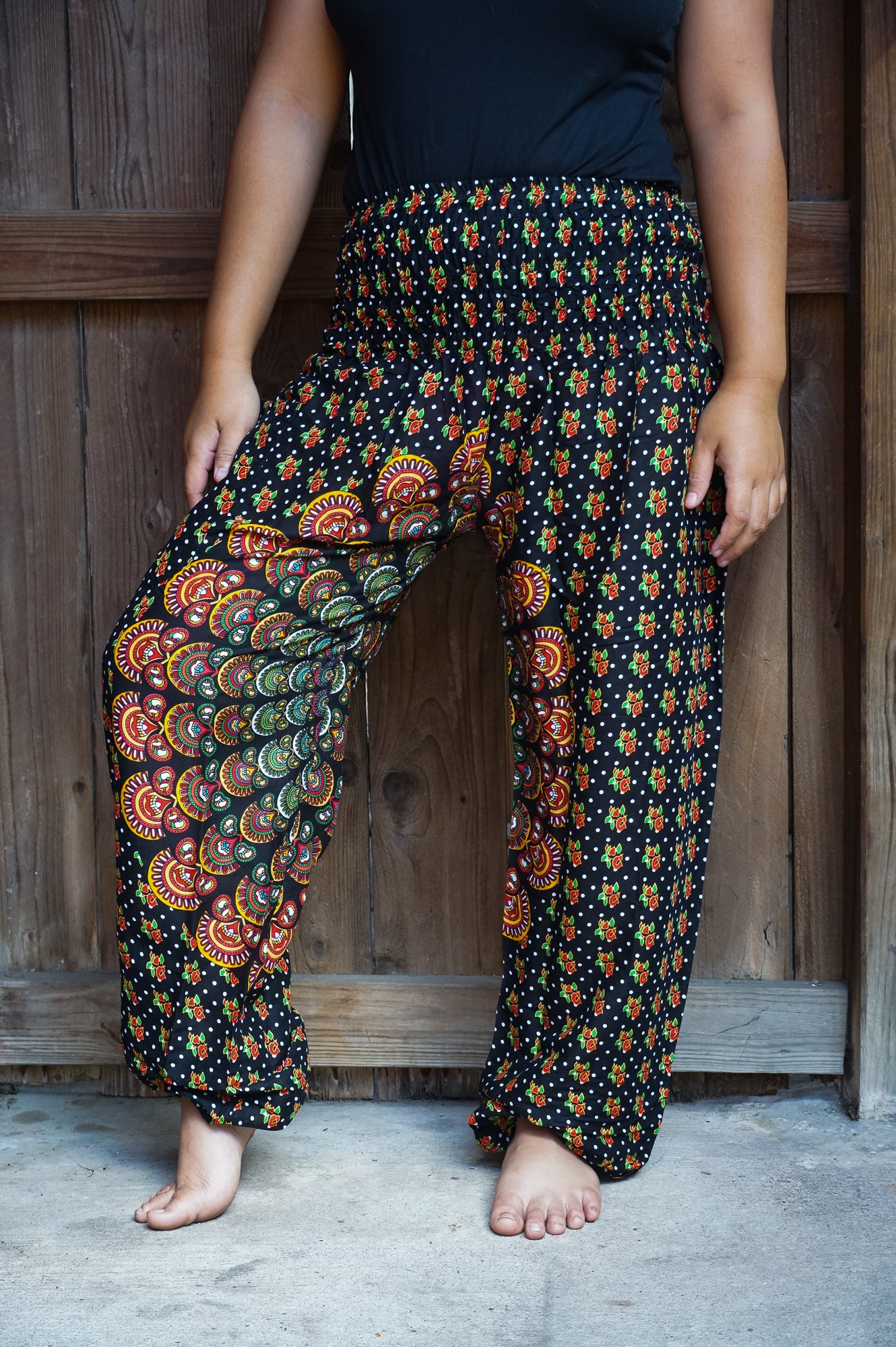 A person stands barefoot, wearing a unisex sleeveless black top and the brightly patterned, colorful high-waisted "Black Chakra Harem Pants, Hippie Pants, Boho Pants" made from 100% Rayon.