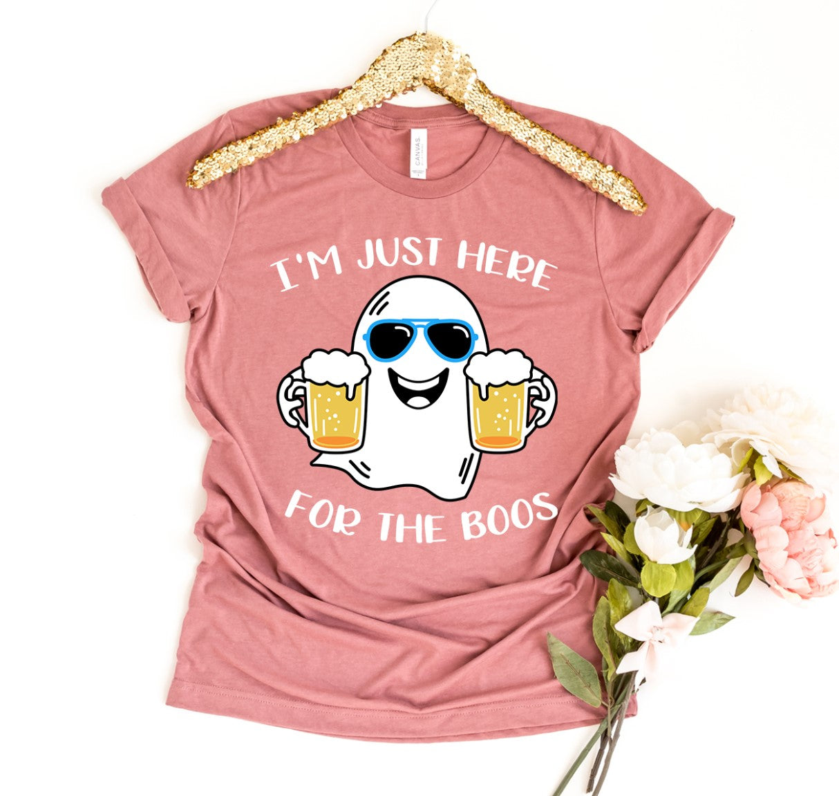 A premium quality, pink "I'm Just Here For The Boos Shirt" featuring a cartoon ghost wearing sunglasses and holding two beer mugs. This comfortable retail fit T-shirt is displayed on a hanger adorned with gold glitter, next to a bouquet of flowers.