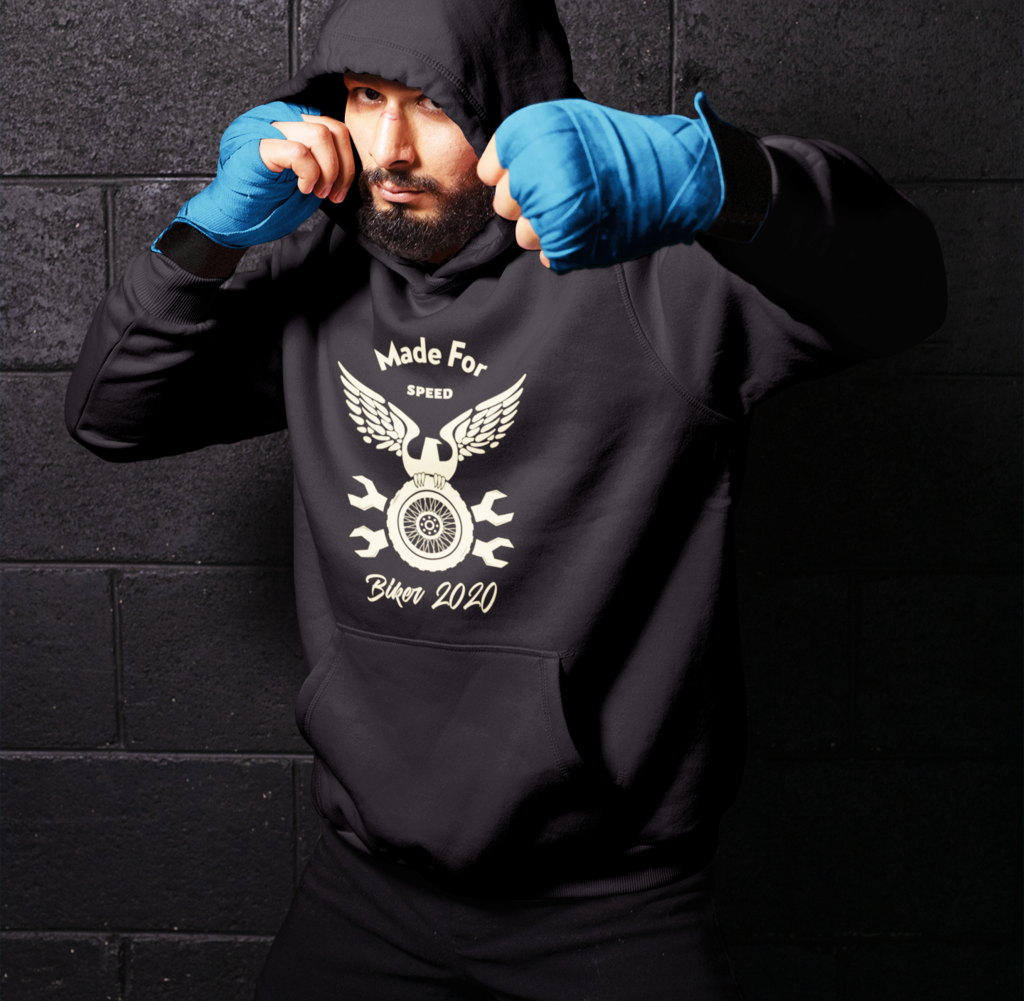 Man in a Made for Speed Hooded Sweatshirt and blue boxing gloves posing in a defensive stance against a dark brick wall.